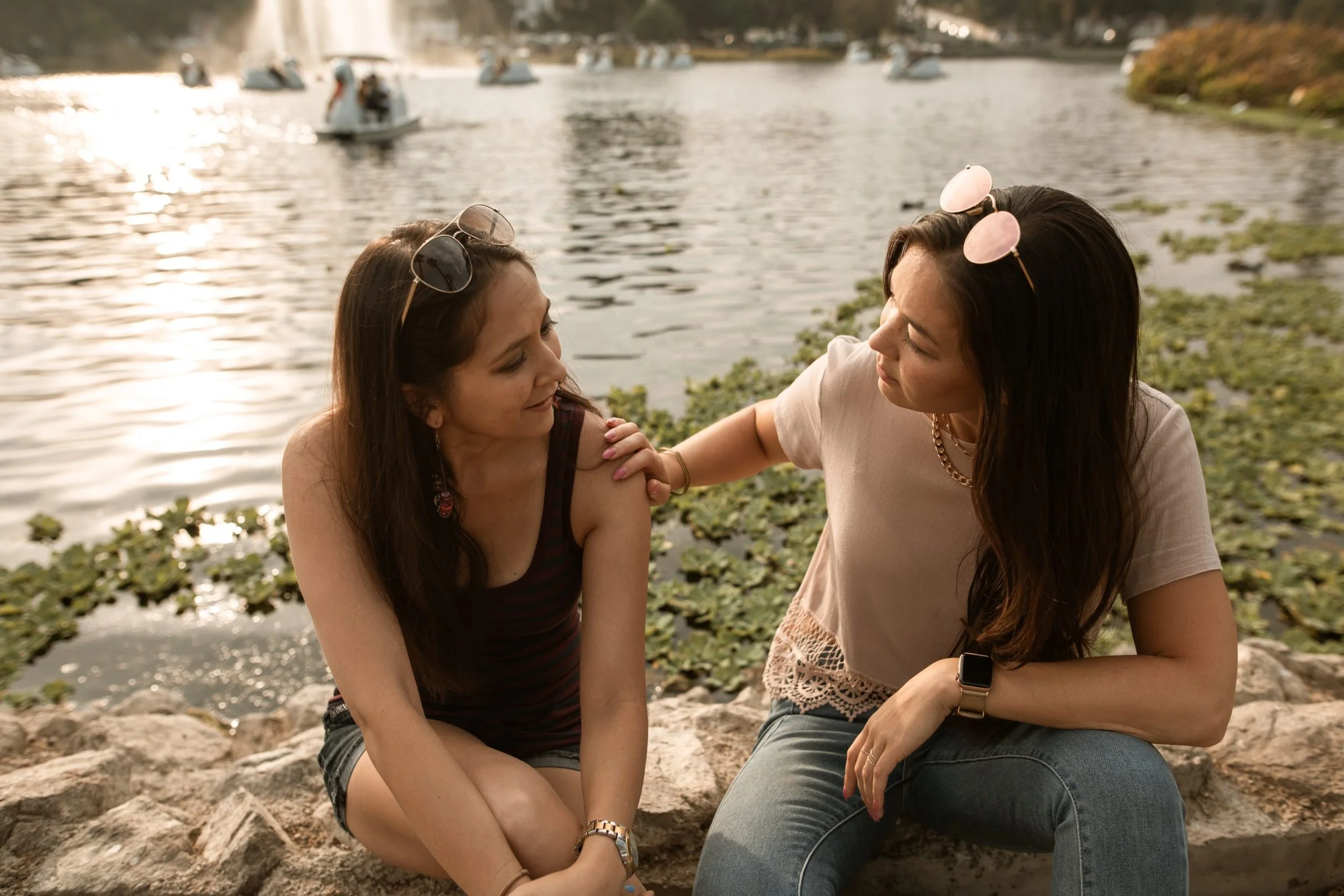Two women sitting on rocks by a lake, one touching the other's shoulder, with paddle boats on the water in the background.