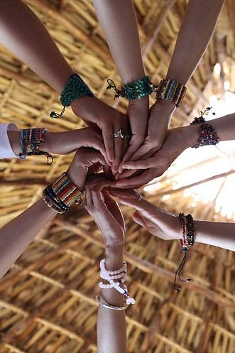 Multiple hands with colorful bracelets and rings come together to form a heart shape against a wooden ceiling background, symbolizing unity and friendship.