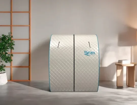 A portable relaxation pod with a quilted exterior, labeled 'Relax', standing in a modern room with light gray walls, a wooden side table, and a potted plant.