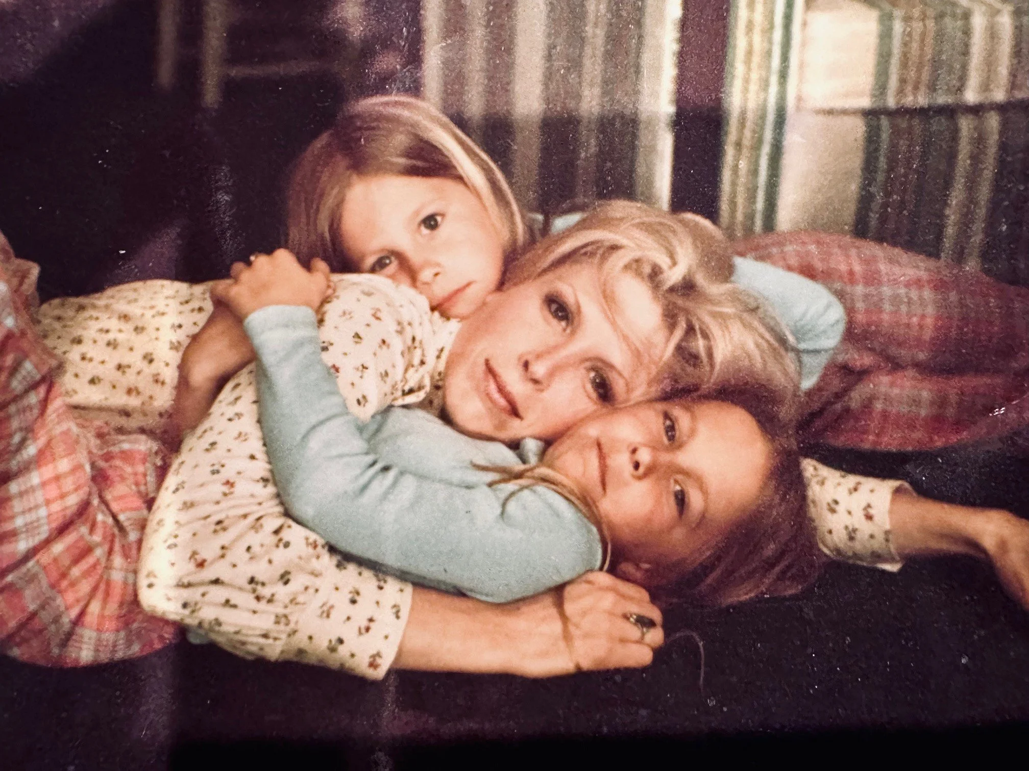 Four people are lying together on a bed, closely embracing and smiling. There are two adults and two children, all with light skin. The background shows a wall with striped wallpaper.