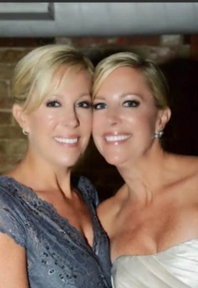 Two women with blonde hair smiling and close together indoors, one in a lace dress and the other in a strapless gown.