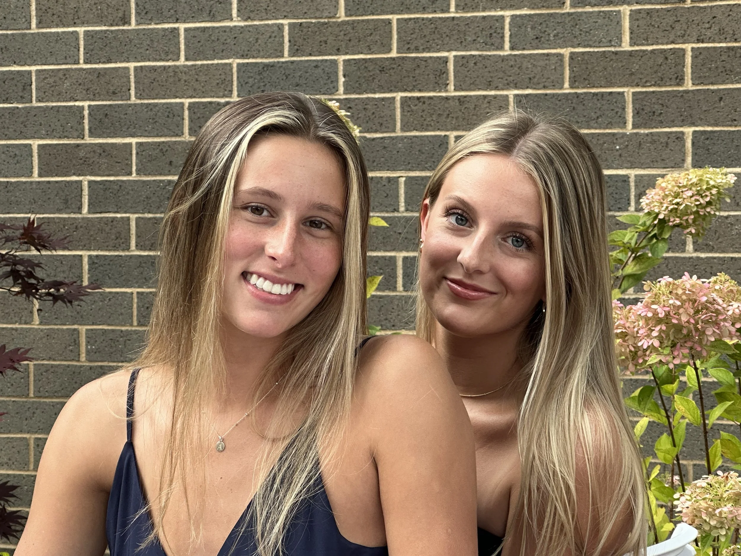 Two young women with long blonde hair smiling, standing outdoors in front of a brick wall and pink hydrangea flowers.