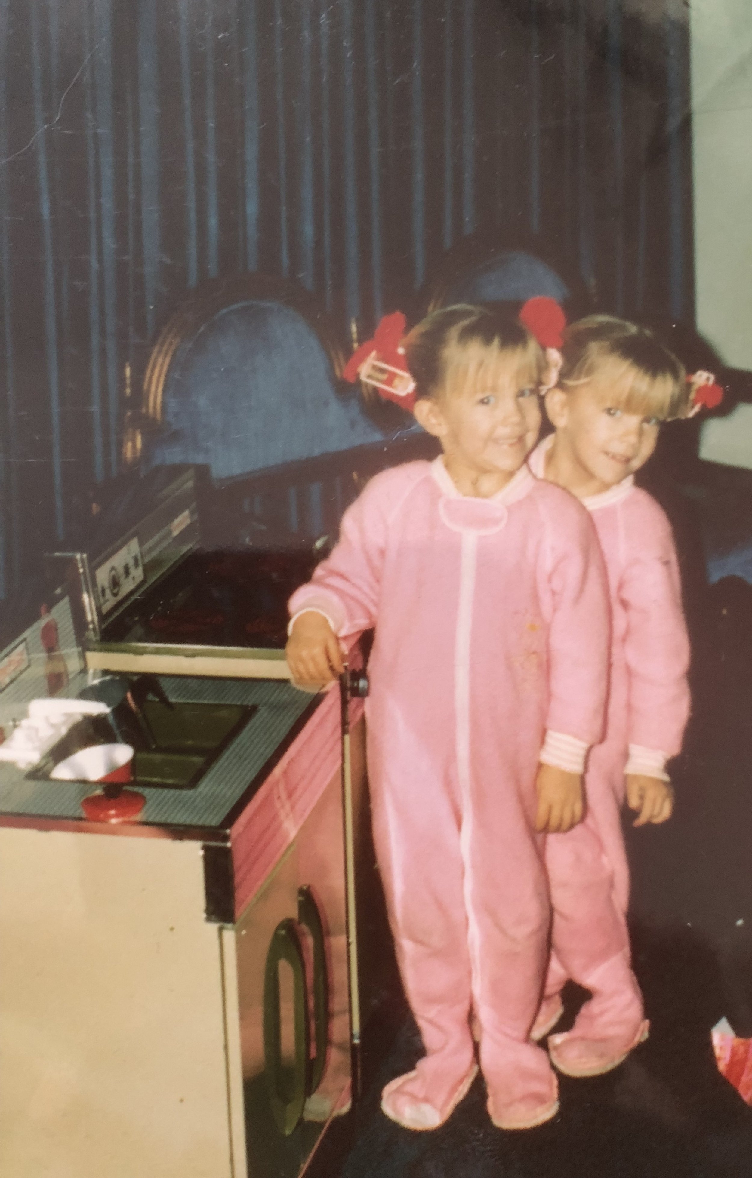 Two young girls with blonde hair wearing pink pajamas standing next to a toy stove, smiling at the camera, with a dark curtain in the background.