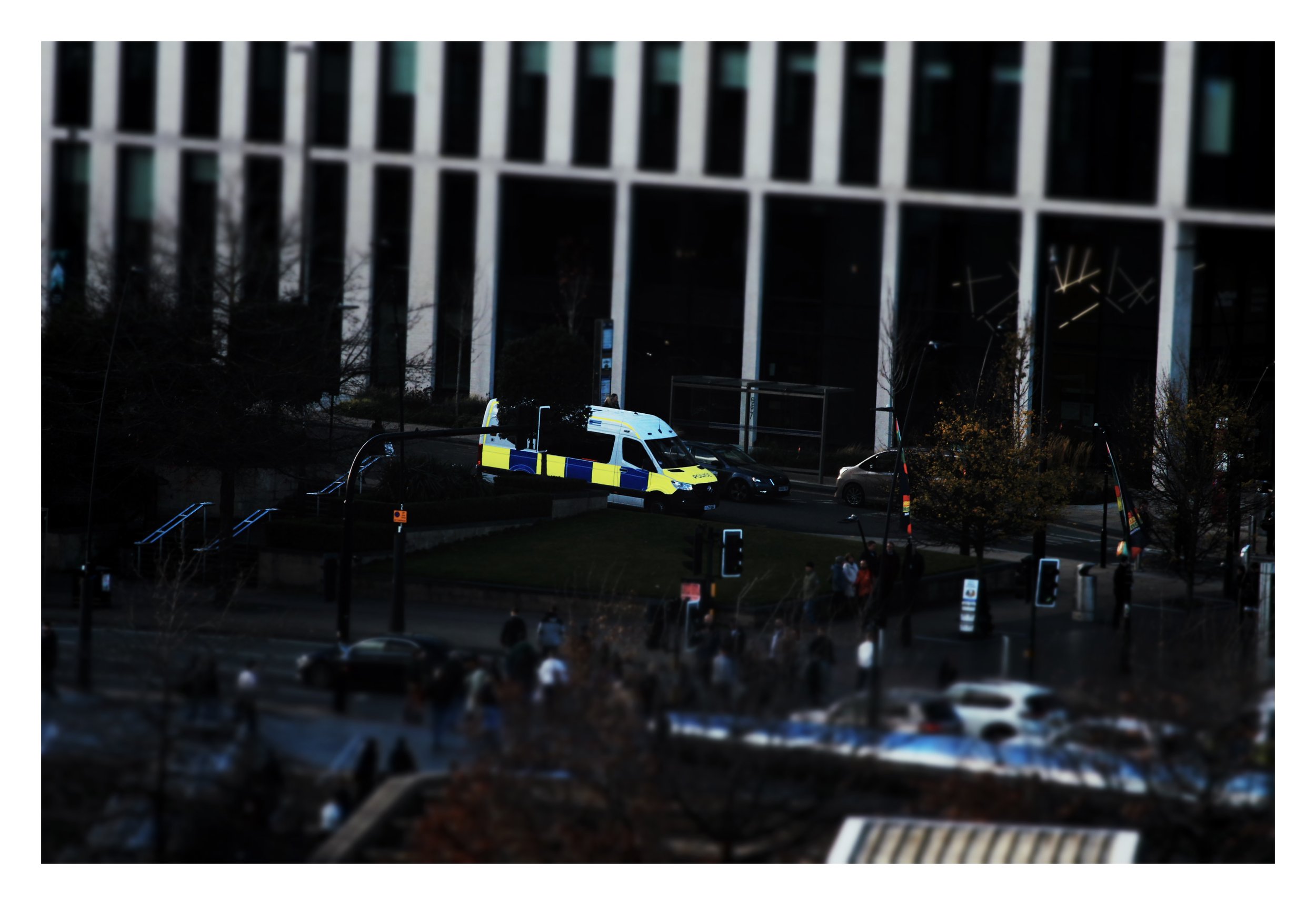City street scene at night with a police van and pedestrians crossing the street in front of a modern building with large columns.