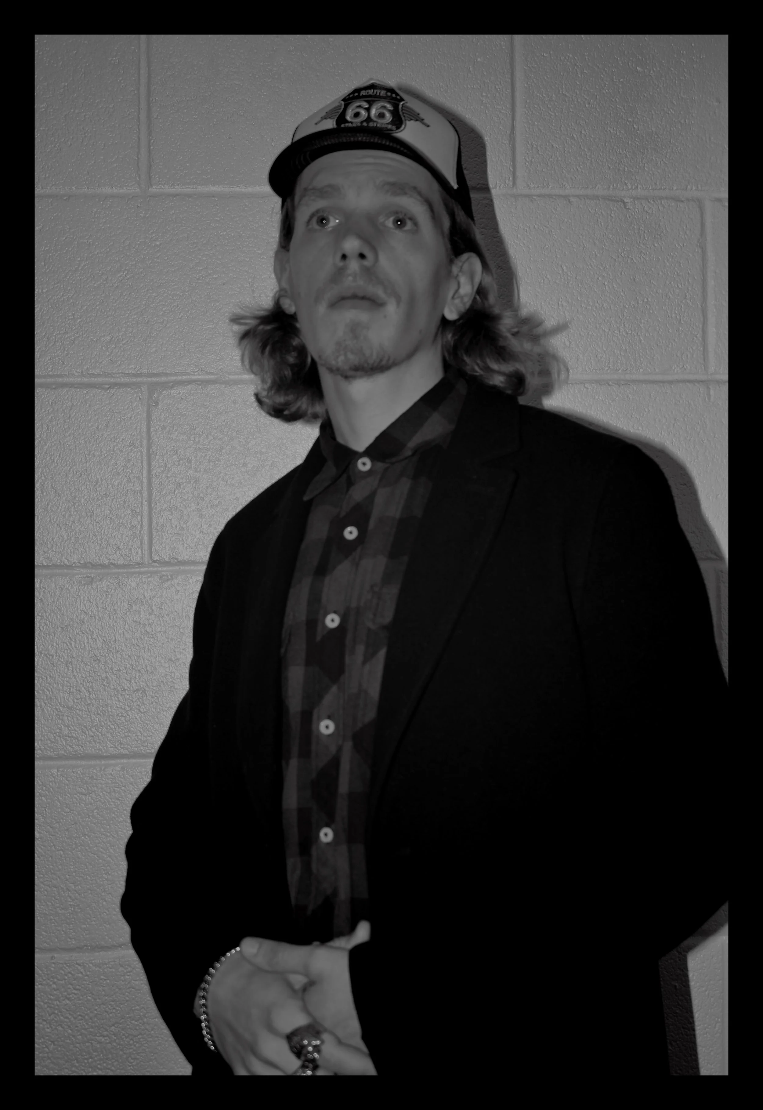 A man with shoulder-length hair, a goatee, and mustache, wearing a cap with a Route 66 logo, a dark blazer, and a checkered shirt, standing against a brick wall.