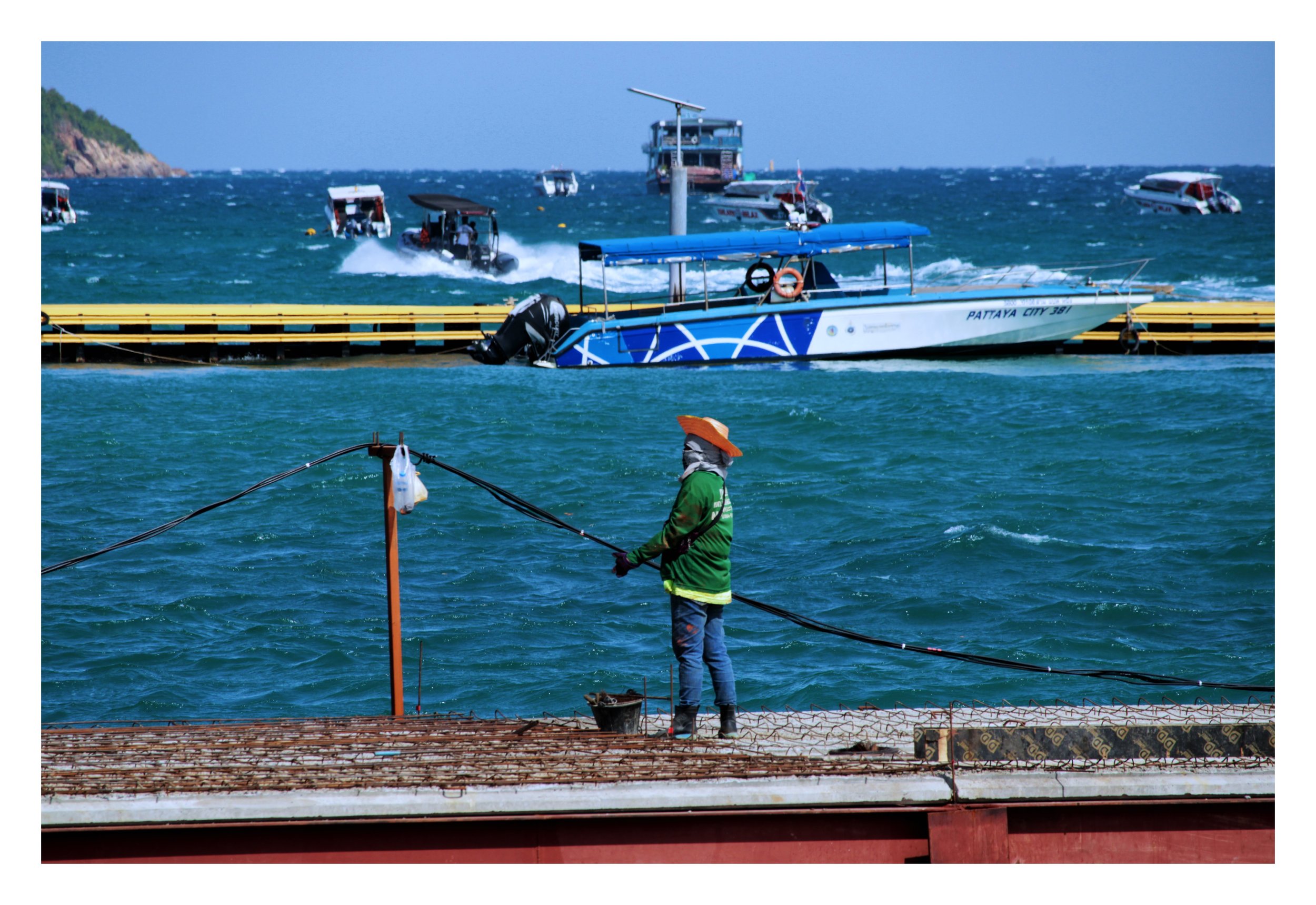 A person fishing off a pier, with boats and water in the background at a busy harbor location.