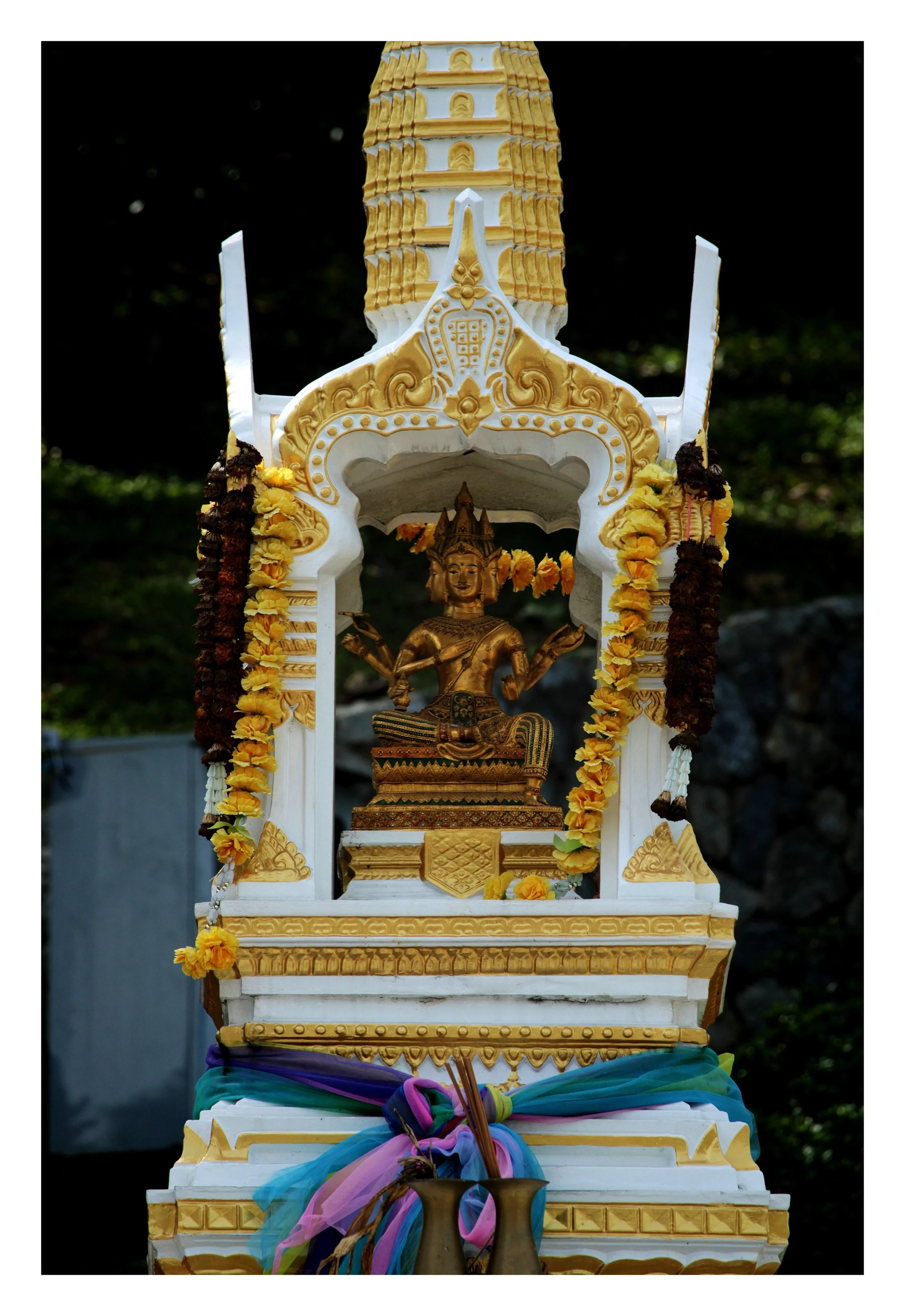 Golden Buddha statue inside an ornate white and gold shrine decorated with flower garlands and colorful fabric, set against a dark outdoor background.