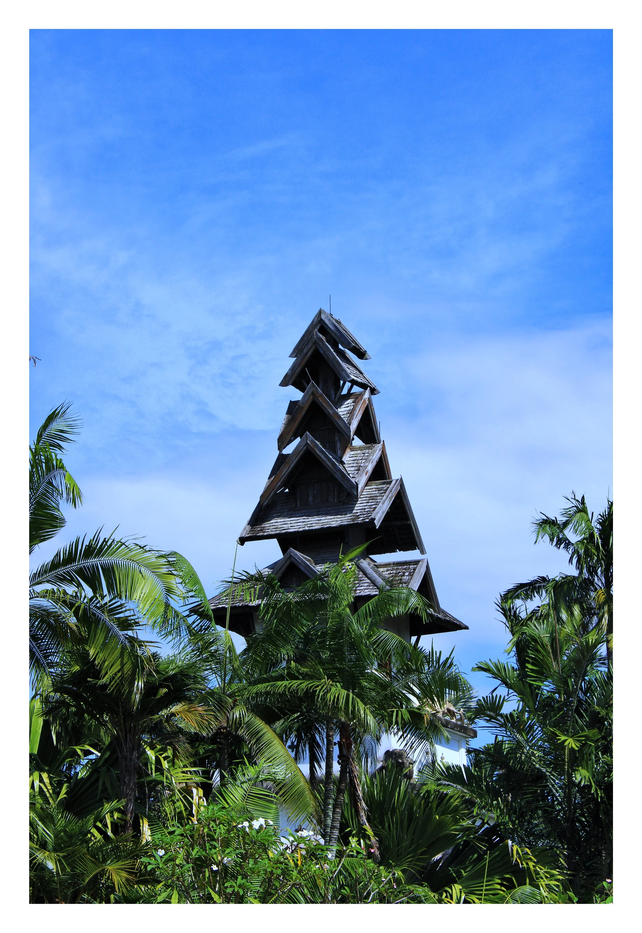A tall wooden structure with multiple small roofs, surrounded by lush green tropical plants and palm trees under a blue sky.