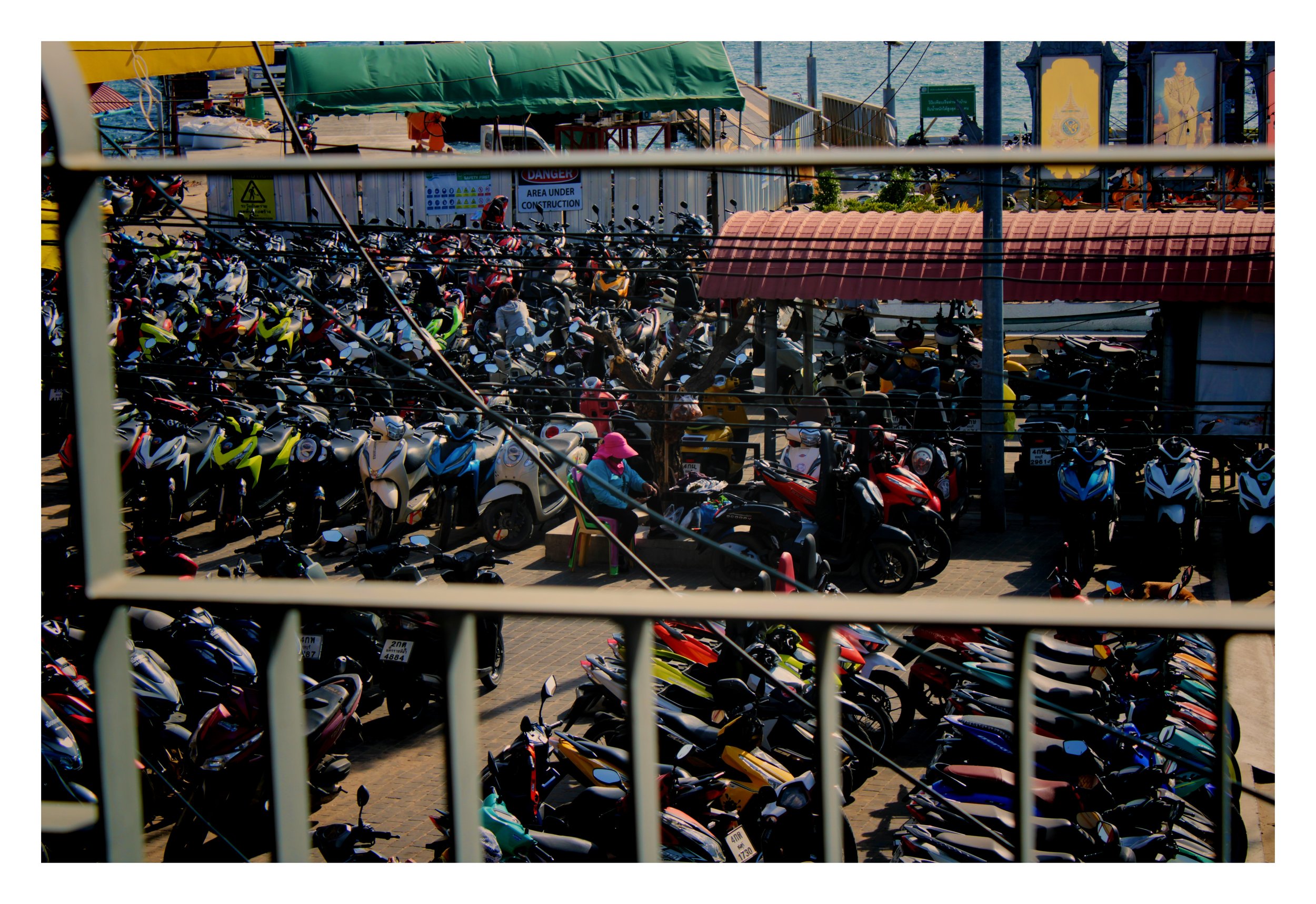 A large parking area filled with many motorbikes and scooters, some parked under shaded covers. The scene is viewed through window bars, with water and a pier visible in the background.
