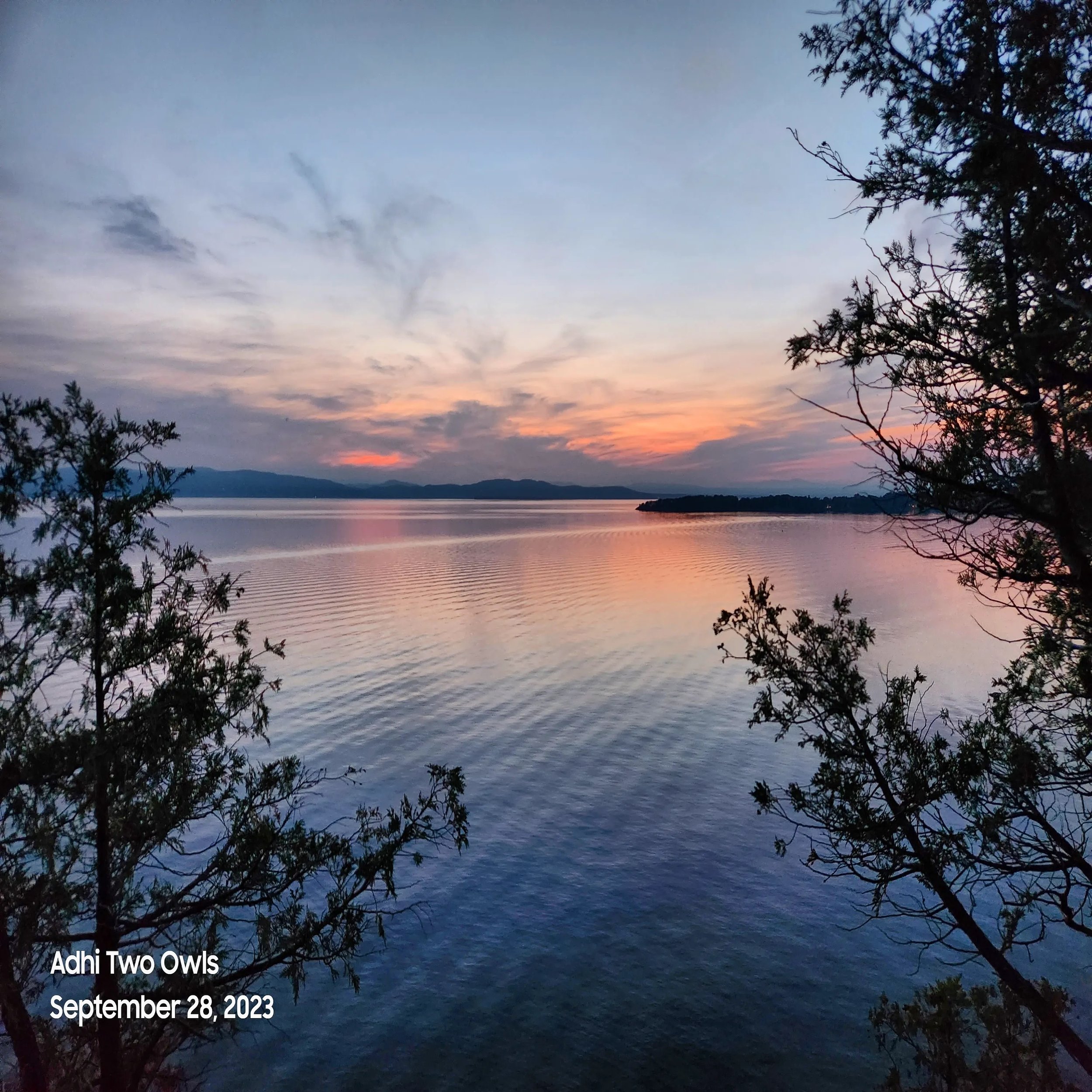 A serene lakeside scene during sunset with calm water reflecting the colorful sky. Trees frame the foreground, and distant mountains are visible on the horizon.