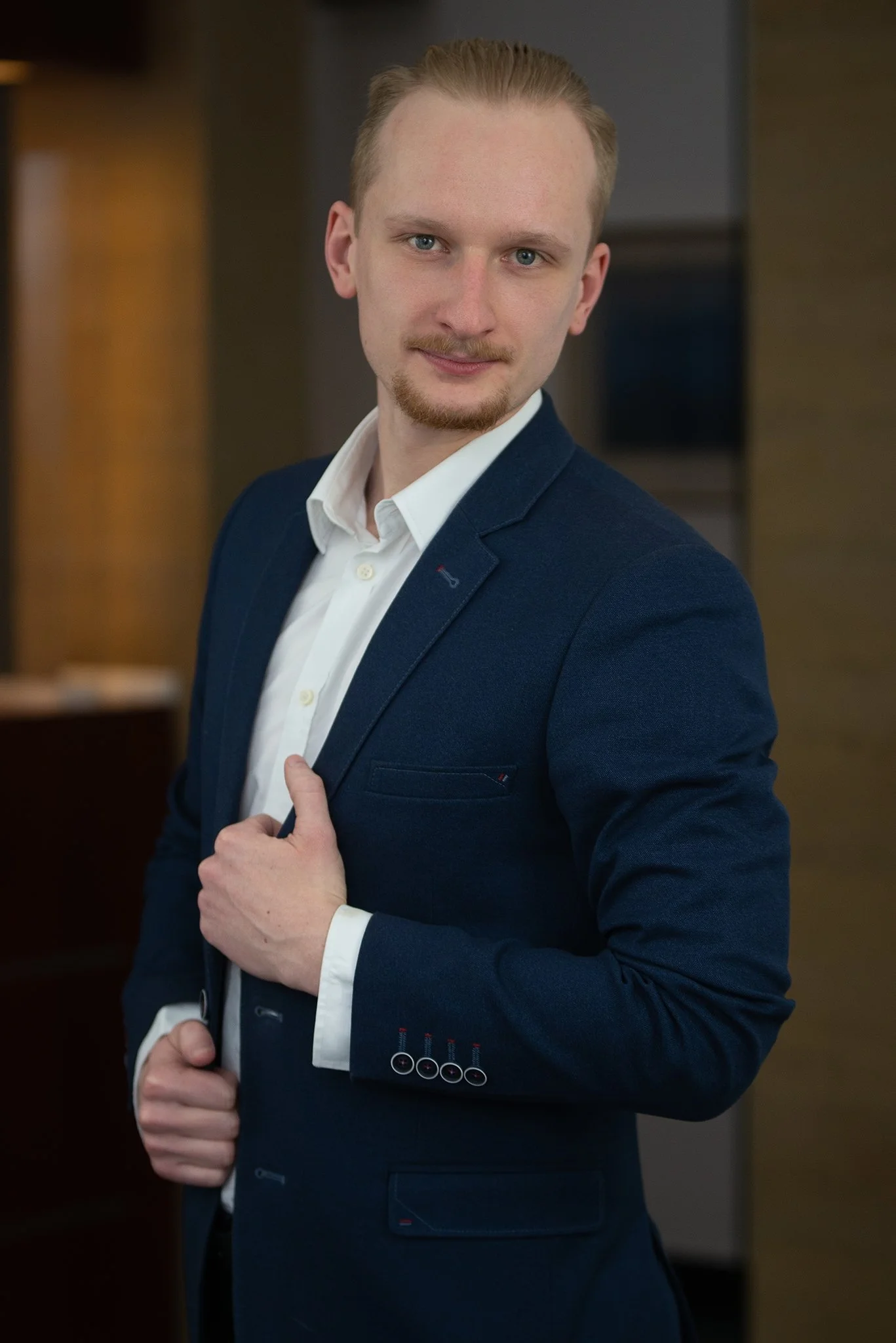 A young man with blond hair and a goatee, wearing a navy blue suit and white shirt, stands indoors with one hand adjusting his jacket and the other arm bent, looking at the camera with a slight smile.