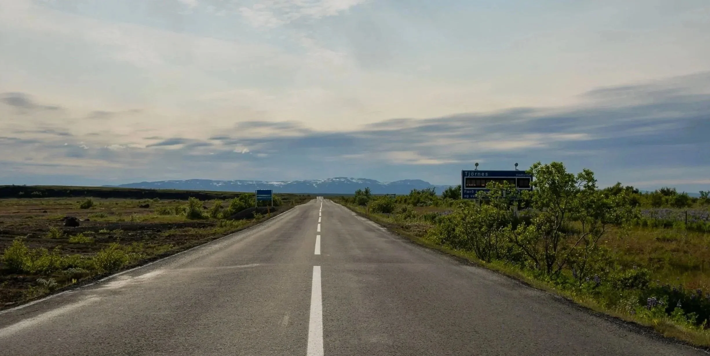 A straight open road in a rural landscape with green bushes and shrubs on both sides, distant mountains with snow-capped peaks under a partly cloudy sky.