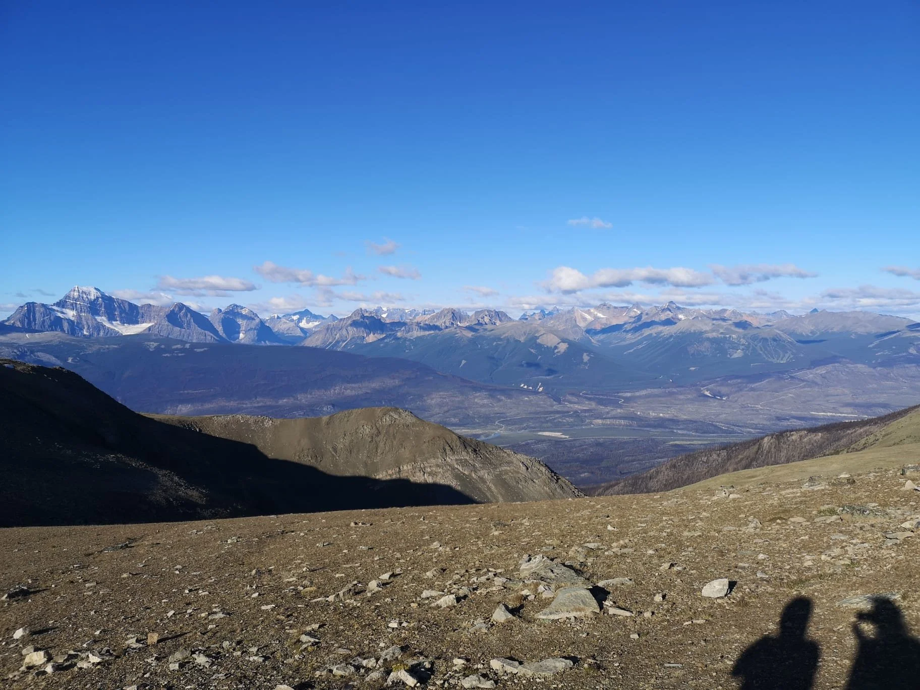 A mountainous landscape with snow-capped peaks under a clear blue sky, taken from a rocky, barren foreground.