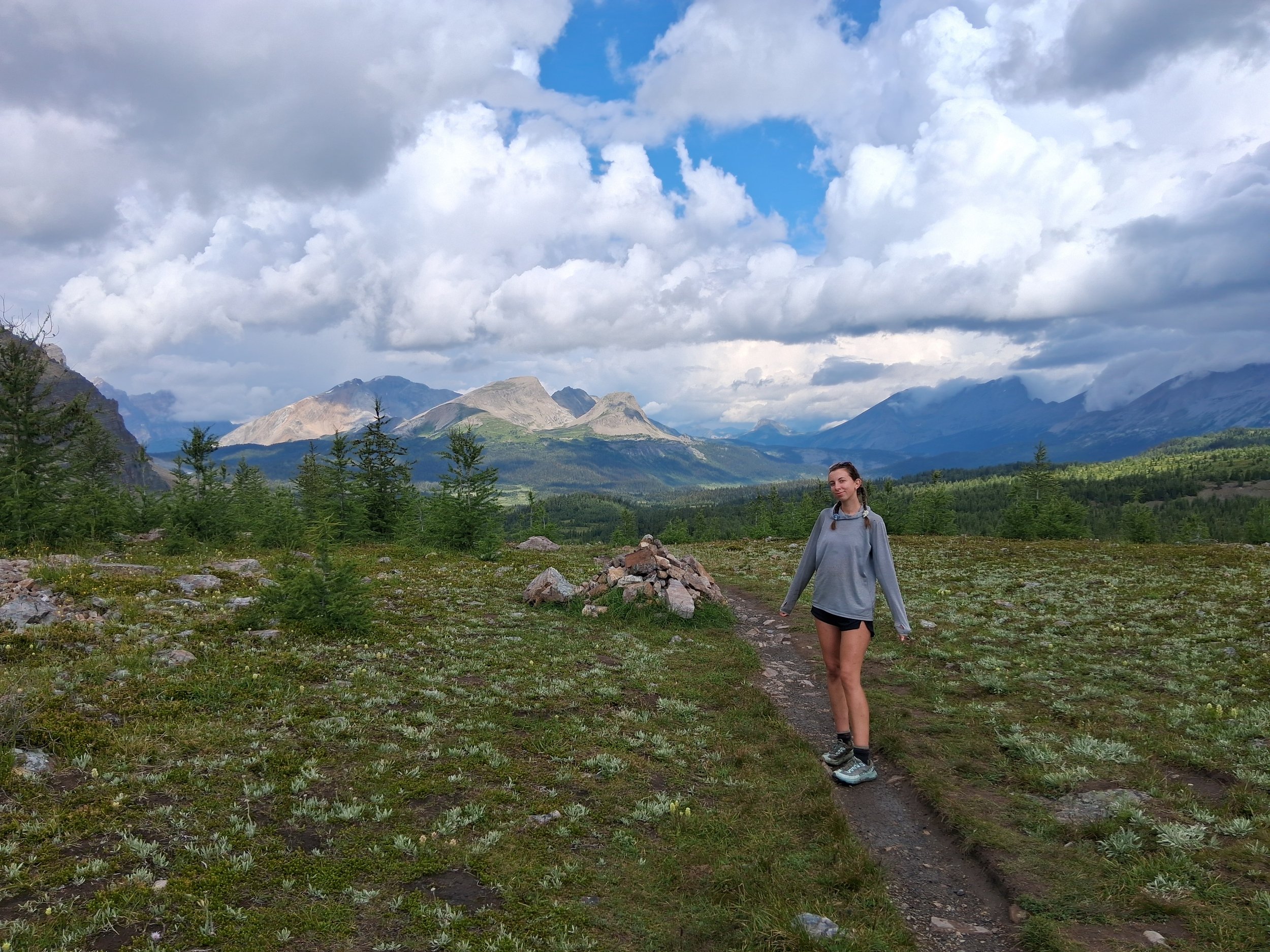 A girl walking on a dirt trail through a grassy field with small trees, mountains in the background, and a cloudy sky.