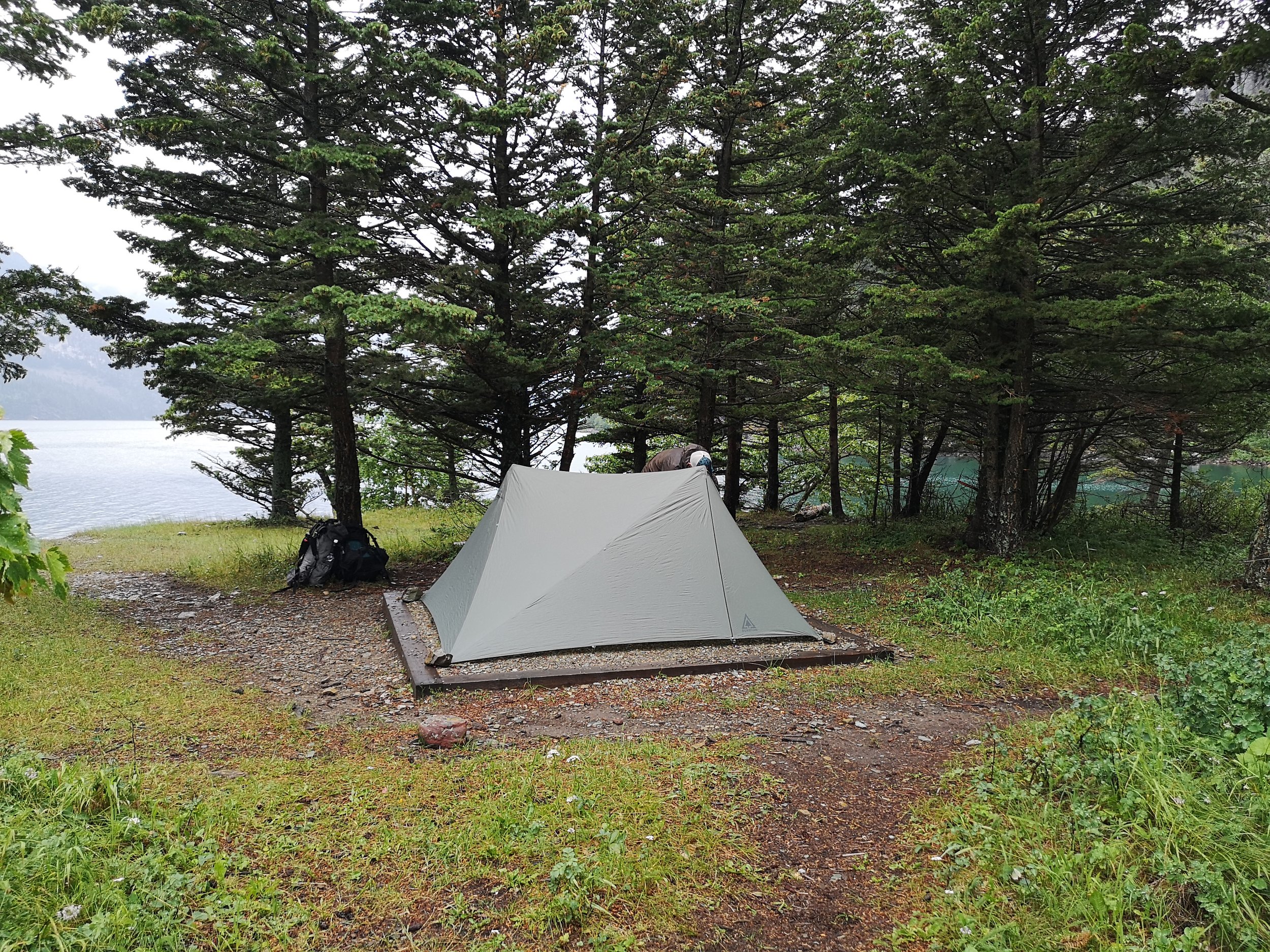 A camping scene with a white tent set up on grass near a lakeside, surrounded by trees
