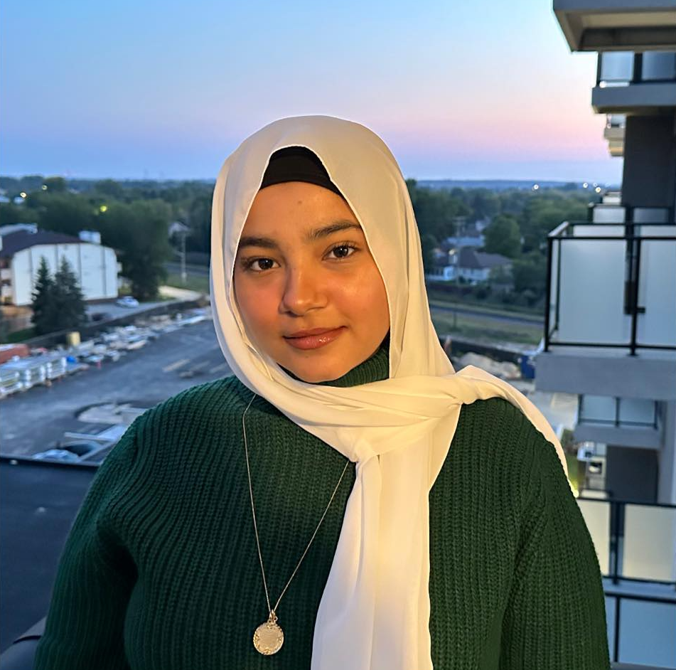 A young woman wearing a white hijab and green sweater standing on a balcony with a cityscape and trees in the background during sunset.