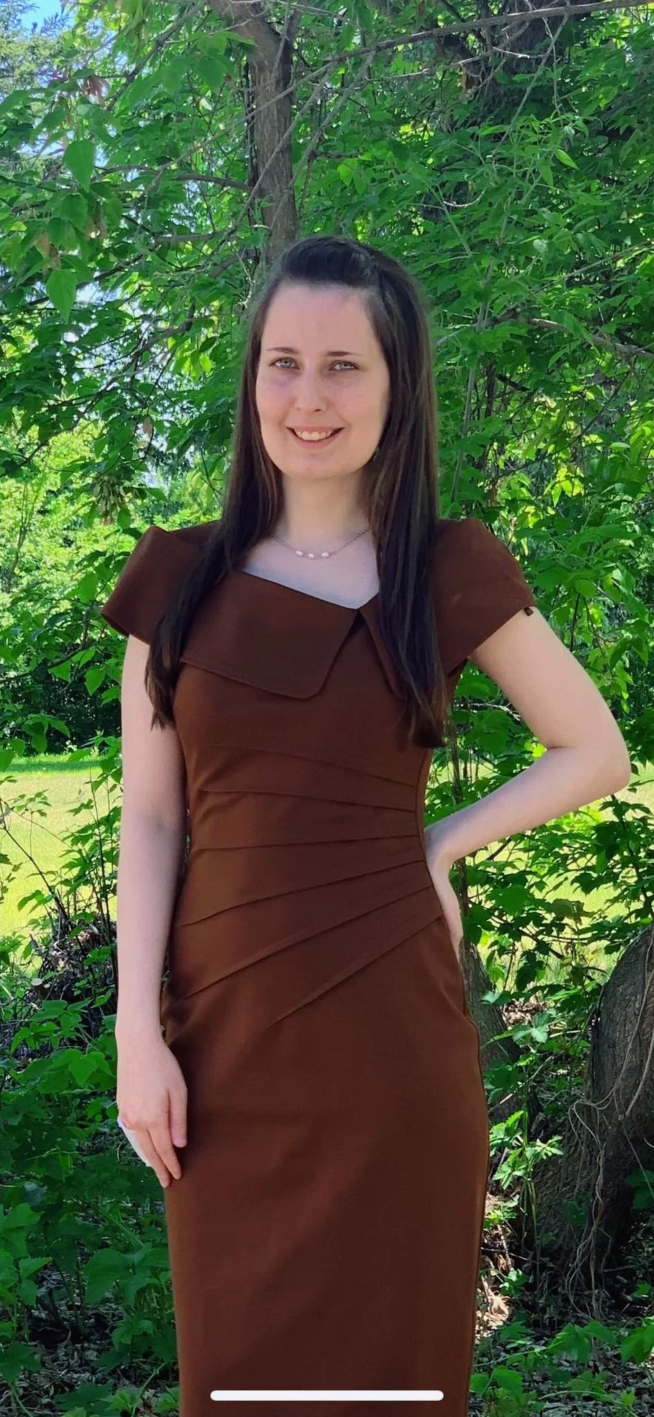 A young woman with long dark hair, wearing a brown dress and a pearl necklace, stands outdoors in front of a green leafy background.