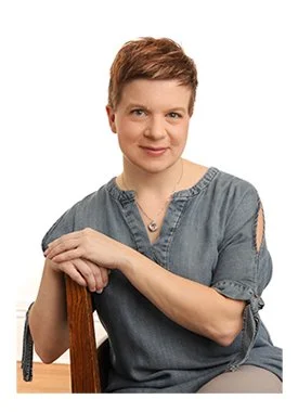 Portrait of a woman with short hair, wearing a gray shirt, sitting on a chair with a white background.