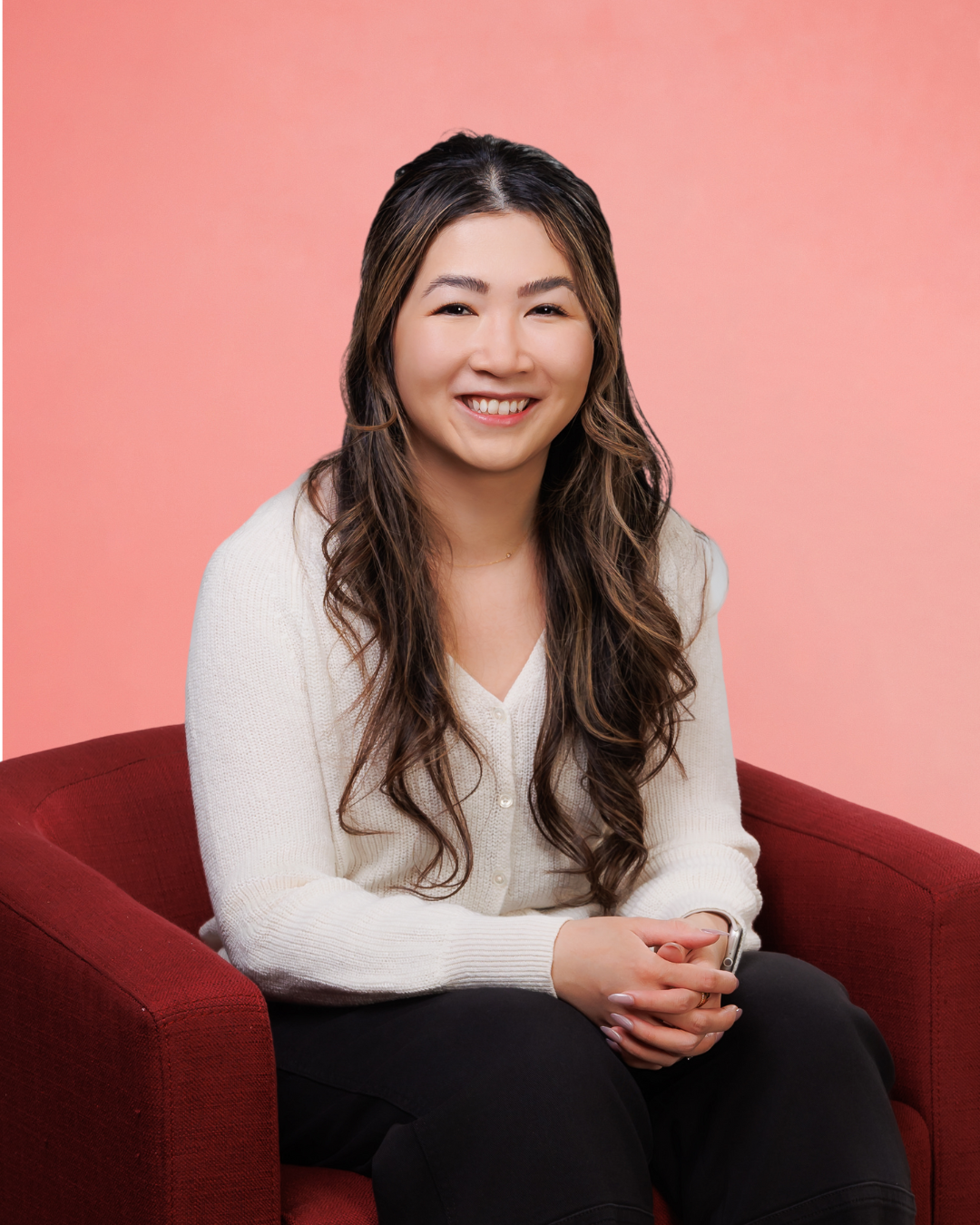 A woman with long wavy hair smiling, sitting on a red armchair against a pink background, wearing a white sweater and black pants.
