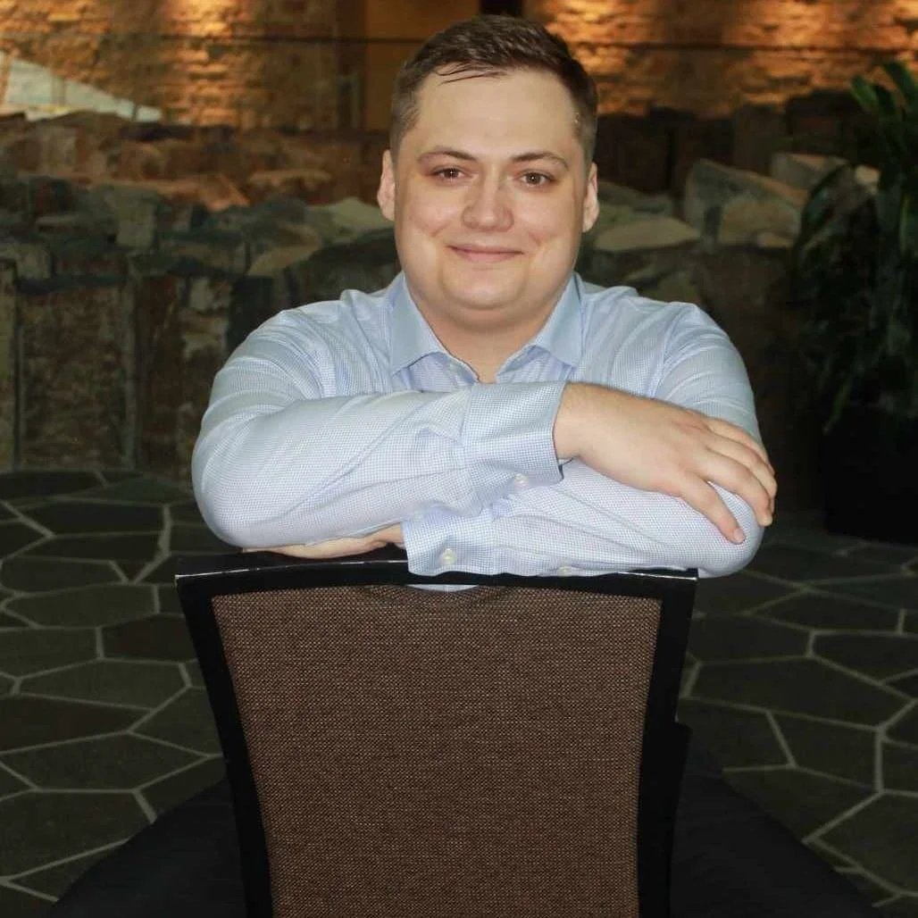 A man with short brown hair and a light blue dress shirt sitting at a table with arms crossed, smiling, in an indoor setting with a rustic brick wall and a stone fireplace in the background.