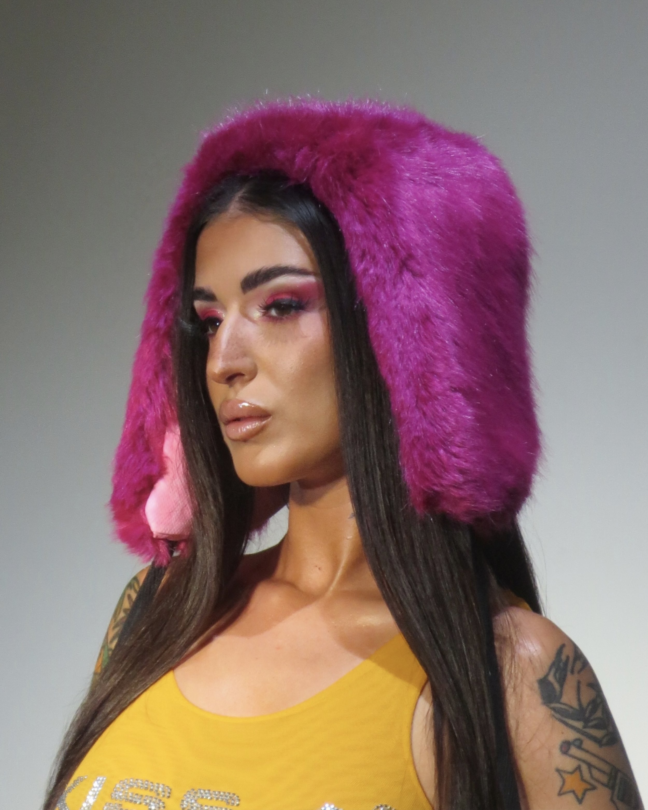 A woman with long dark hair wearing a bright pink furry hood with pink earmuffs, a yellow tank top, and makeup with pink eyeshadow.