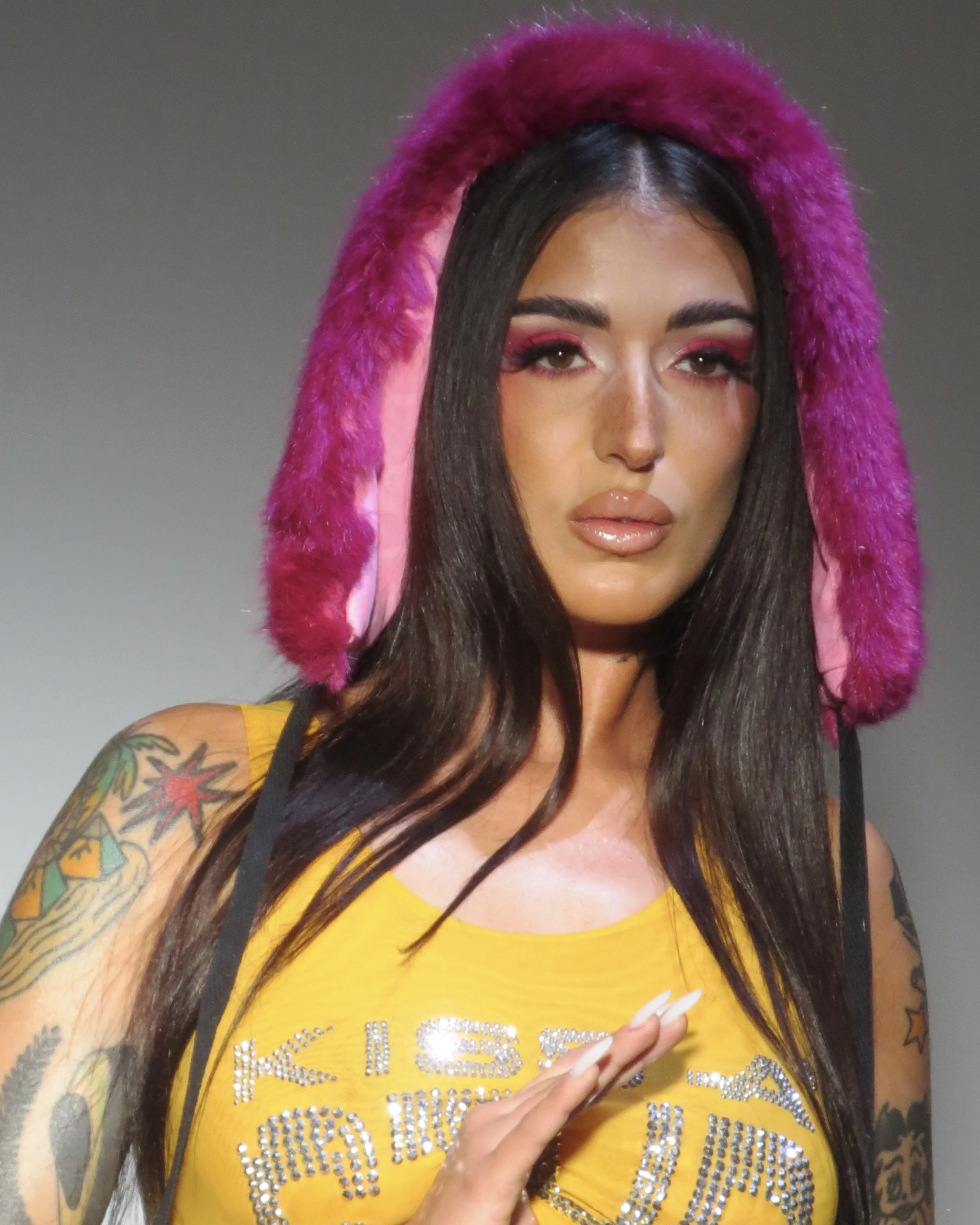 A woman with long dark hair, tattoos on her arms, wearing a yellow top with embellishments, and a pink and purple fur hat.