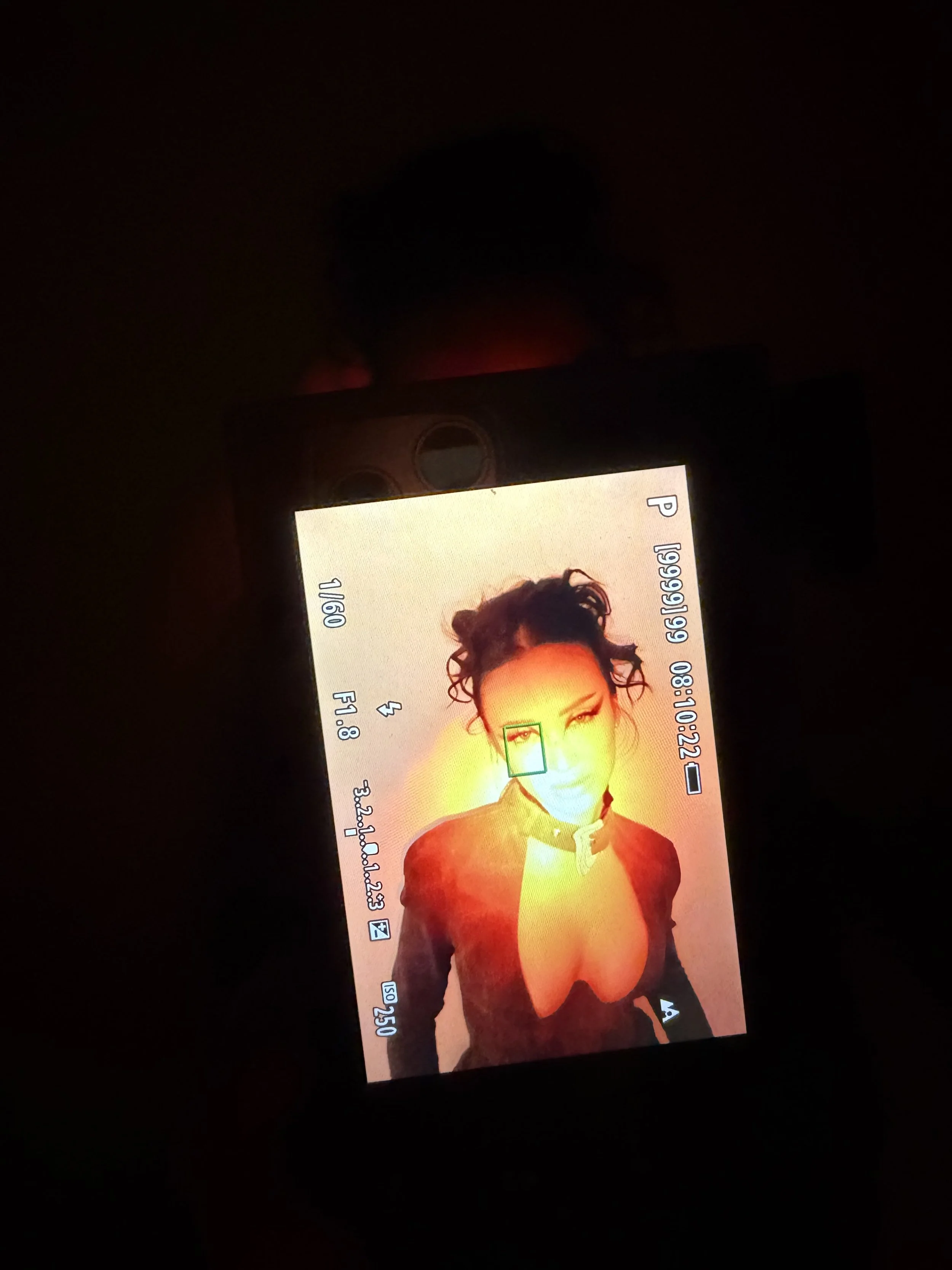 A camera screen displaying a photo of a woman with curly hair, wearing a top with a deep neckline, illuminated by red and orange lighting.