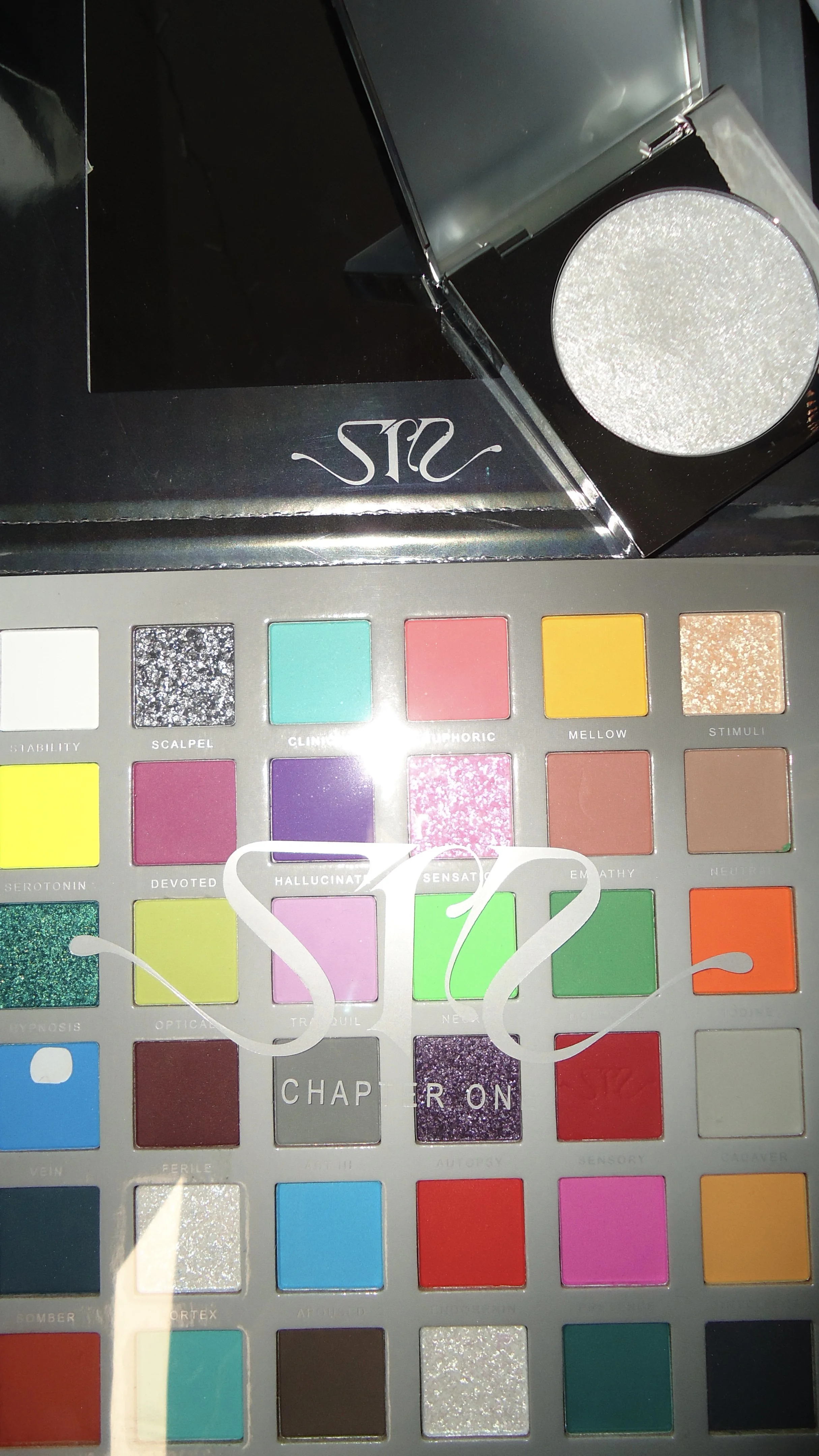 An eyeshadow palette with various colorful shades and a round mirror, with a single eyeshadow pan open showing a shimmering silver color.