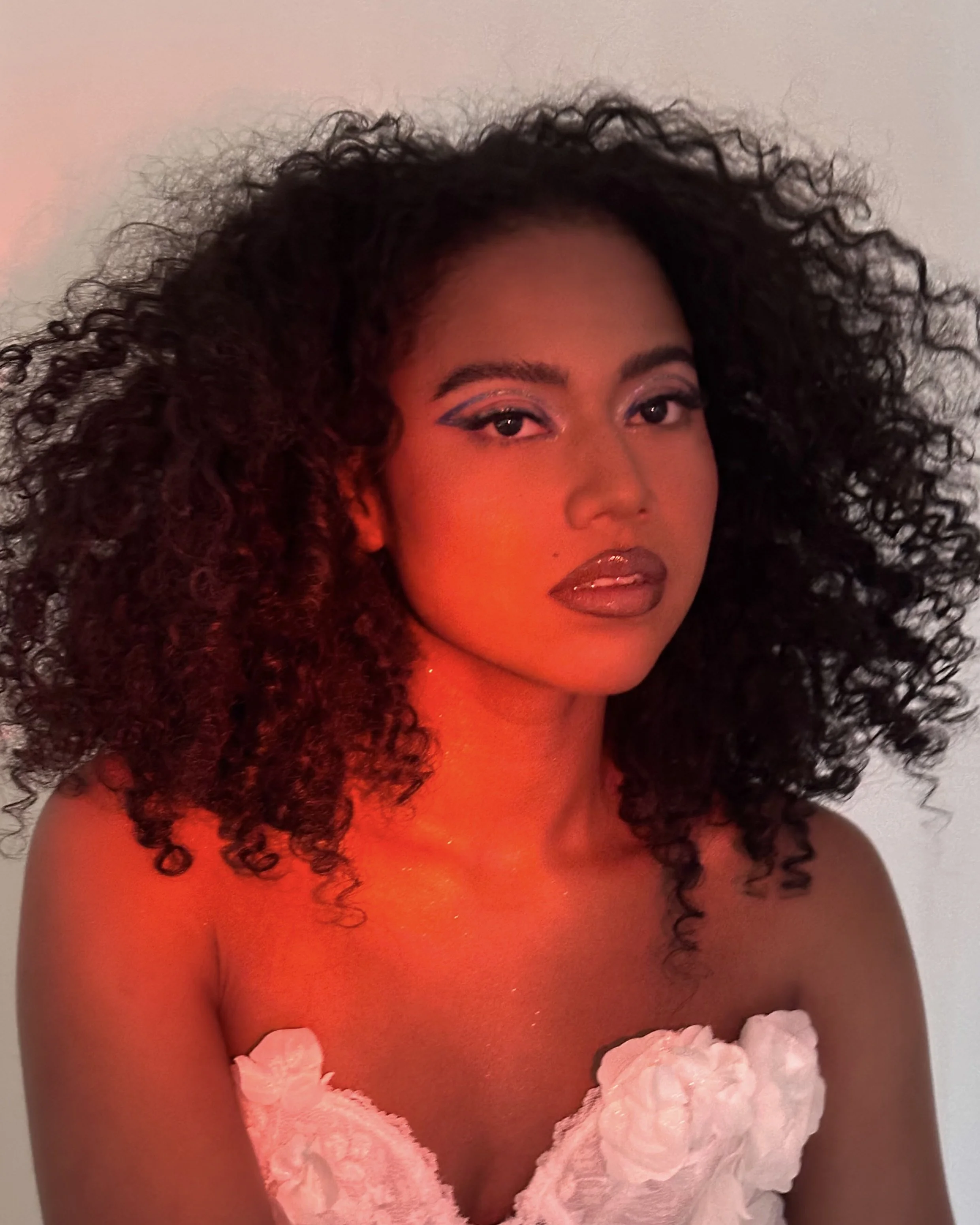 A woman with curly hair and makeup, wearing a strapless white dress with floral details, illuminated by red lighting.