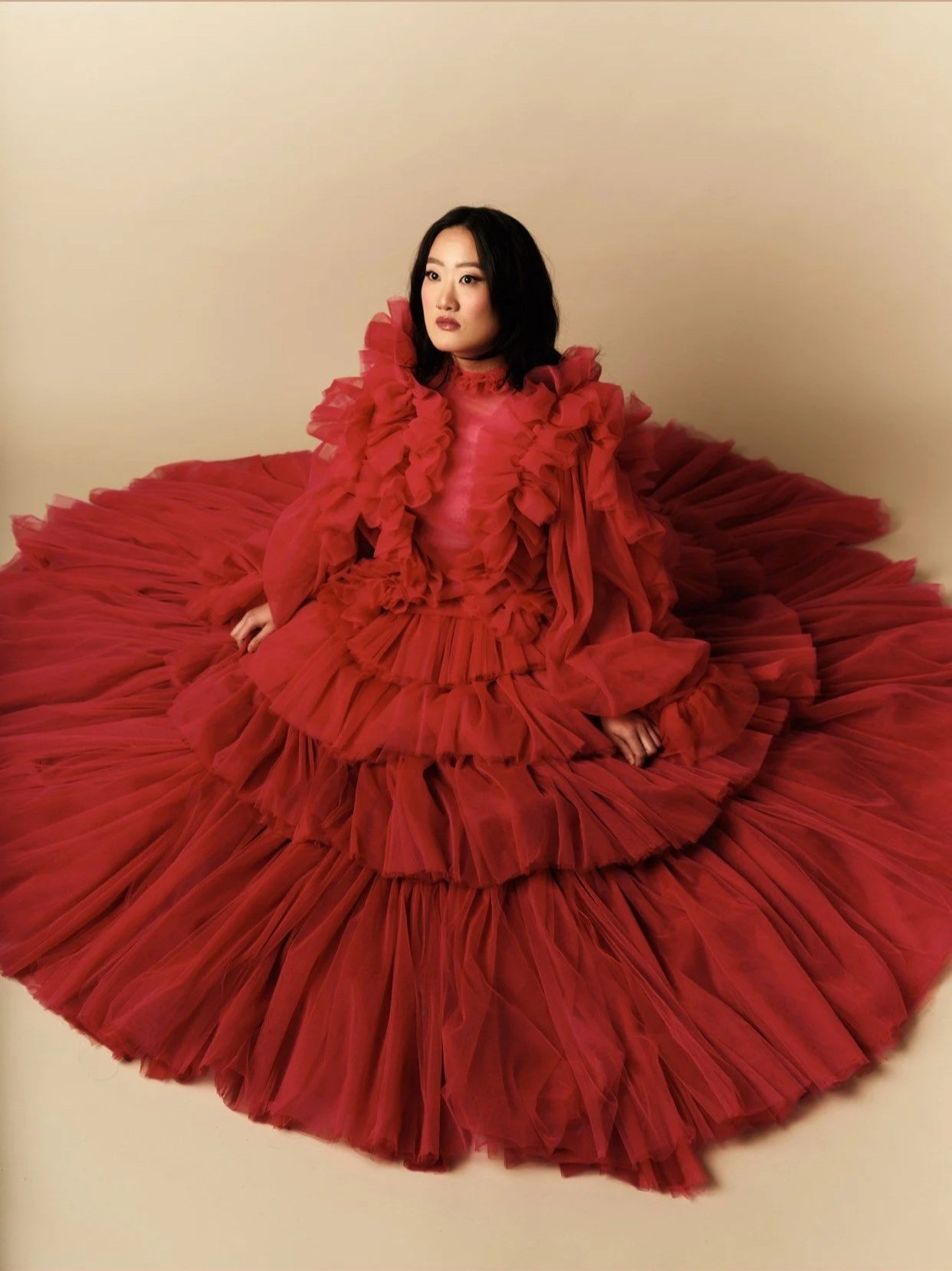 A woman wearing a large, vibrant red, ruffled, floor-length gown sitting on the floor against a plain beige background.