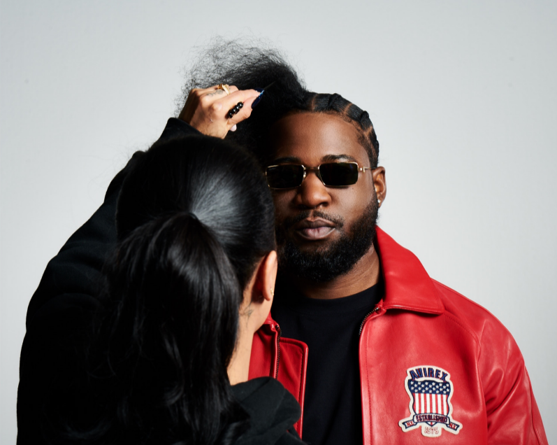 A woman styles a man's hair, who is wearing sunglasses and a red leather jacket with a 'Navirex' patch.