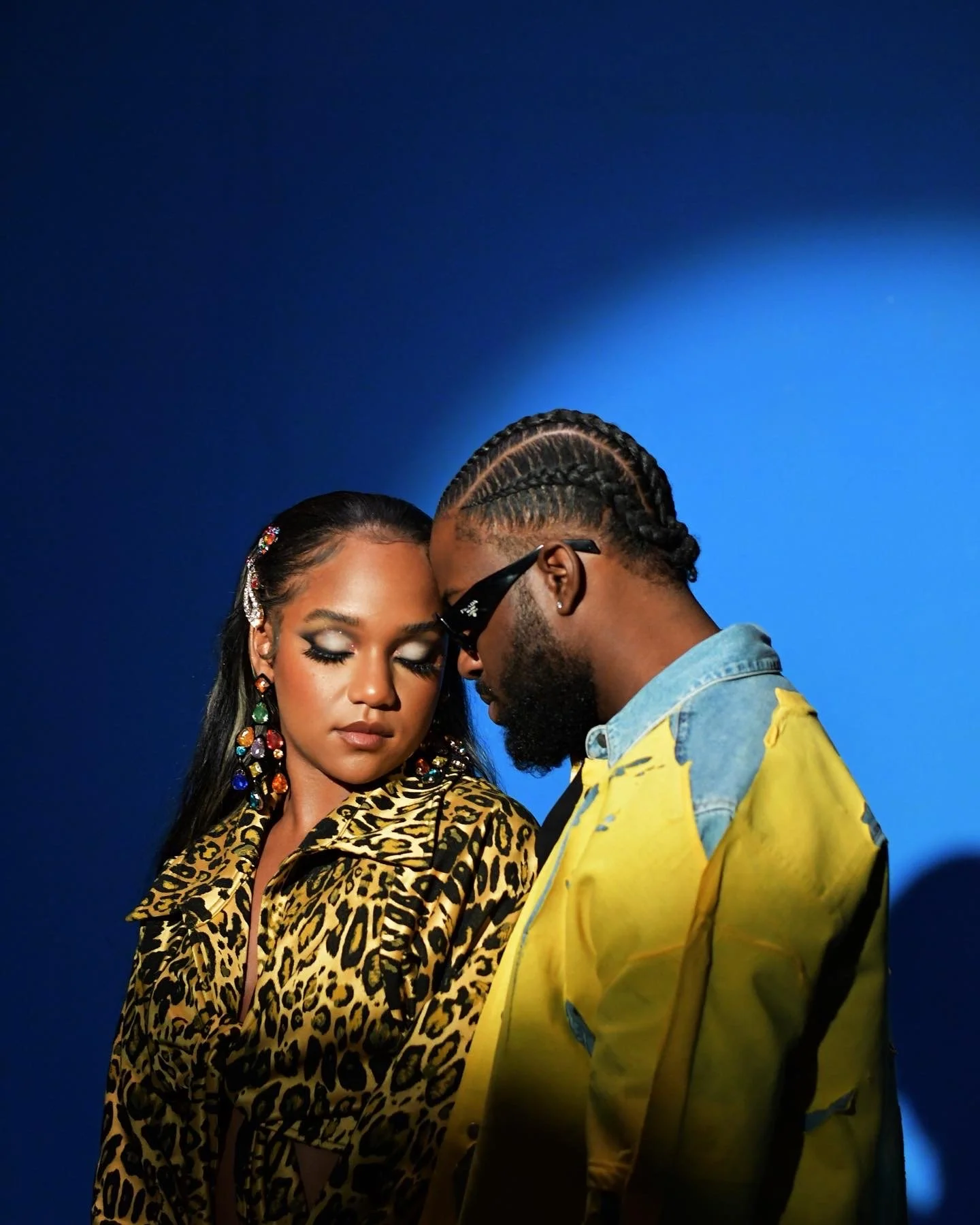 A woman with long dark hair and colorful earrings and makeup, wearing a leopard print jacket, and a man with glasses, braided hair, and a beard, wearing a yellow jacket with blue accents, facing each other with their foreheads touching against a blue