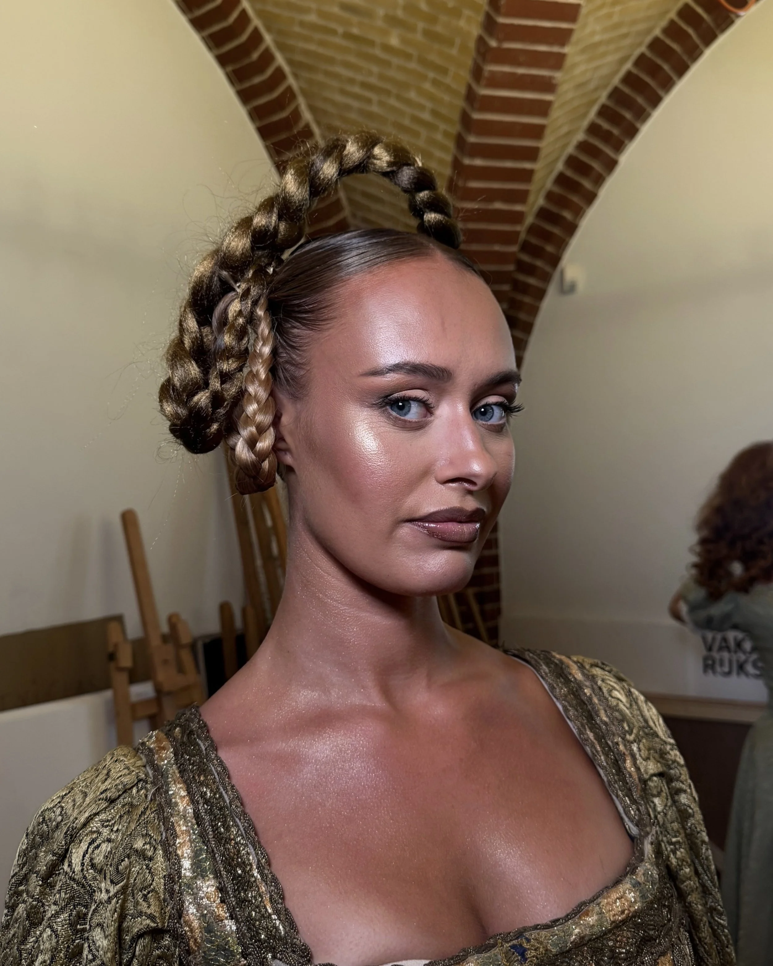 A woman with braided hairstyle, wearing a gold and black patterned garment, standing indoors with a brick arch ceiling.