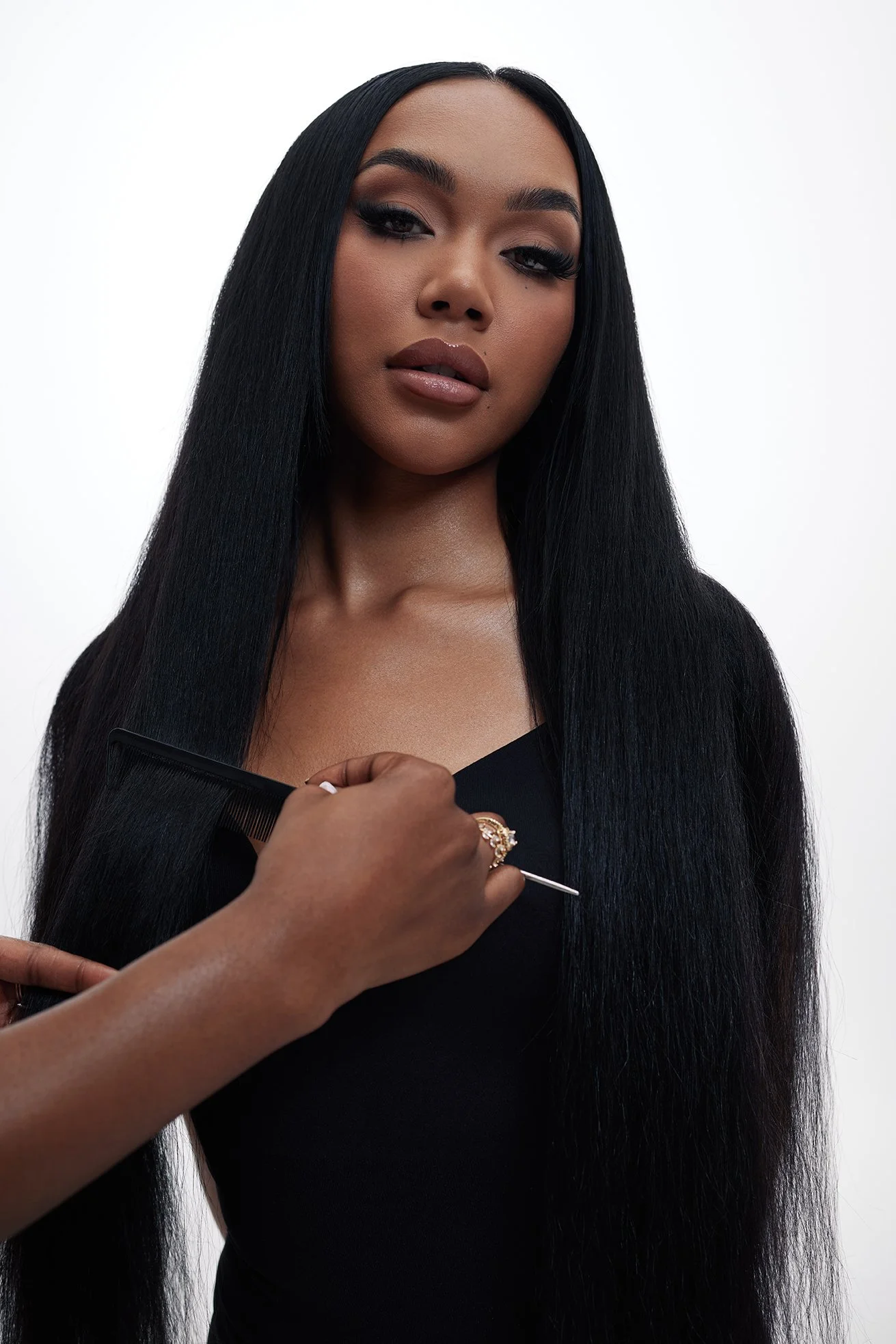 A woman with long black hair being styled or groomed, with makeup, against a plain white background