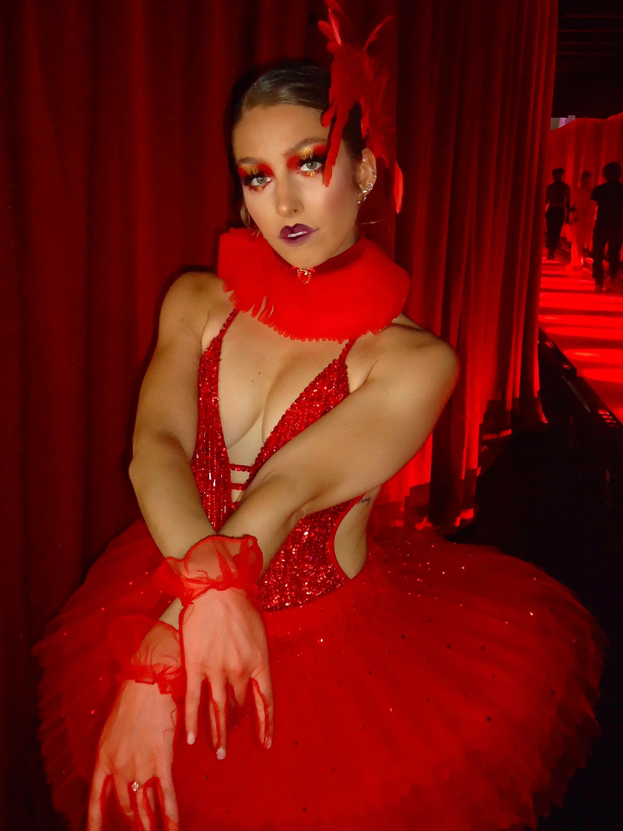 Woman dressed in a red, sequined gown with a full tulle skirt, accessorized with red feathers in her hair and red feathers around her neck, standing in front of red curtains along a red-lit runway.