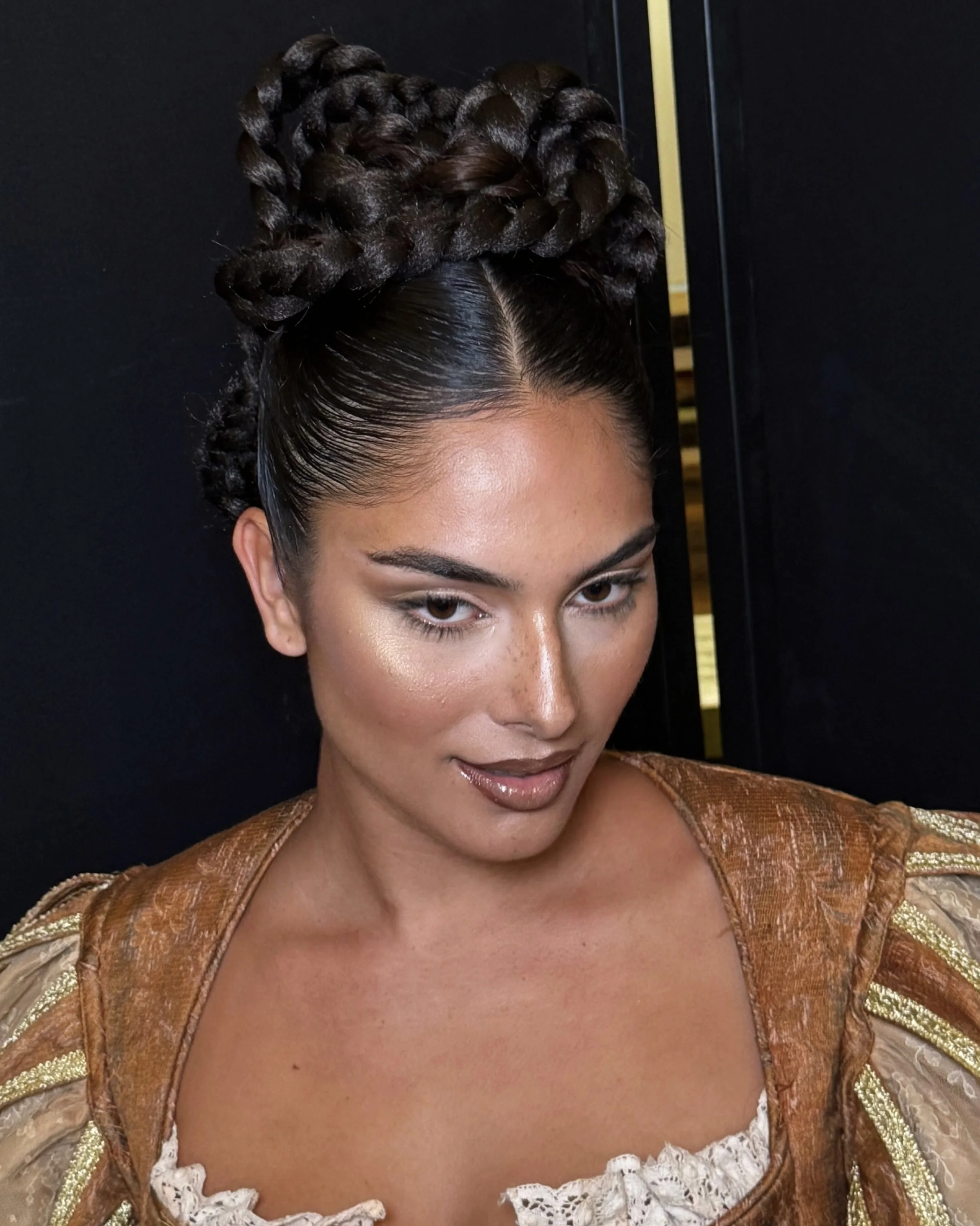 A woman with dark hair styled in intricate braids and twists, wearing a gold and cream dress with lace details and puffed sleeves, looking down with a subtle smile.