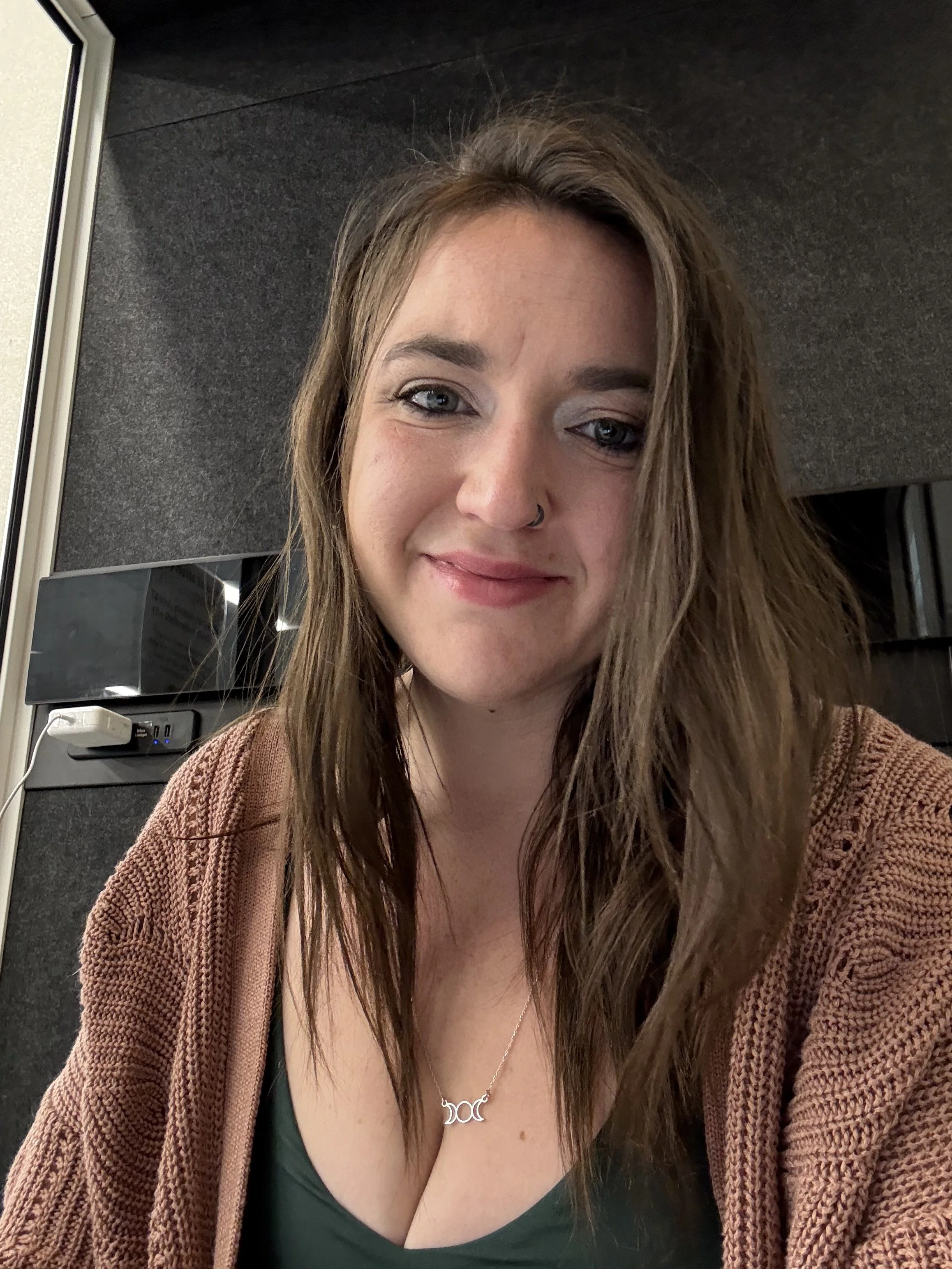 A young woman with long brown hair, blue eyes, and a nose ring smiling while taking a selfie indoors. She's wearing a green top, a brown cardigan, and a silver necklace with a triple crescent moon pendant.