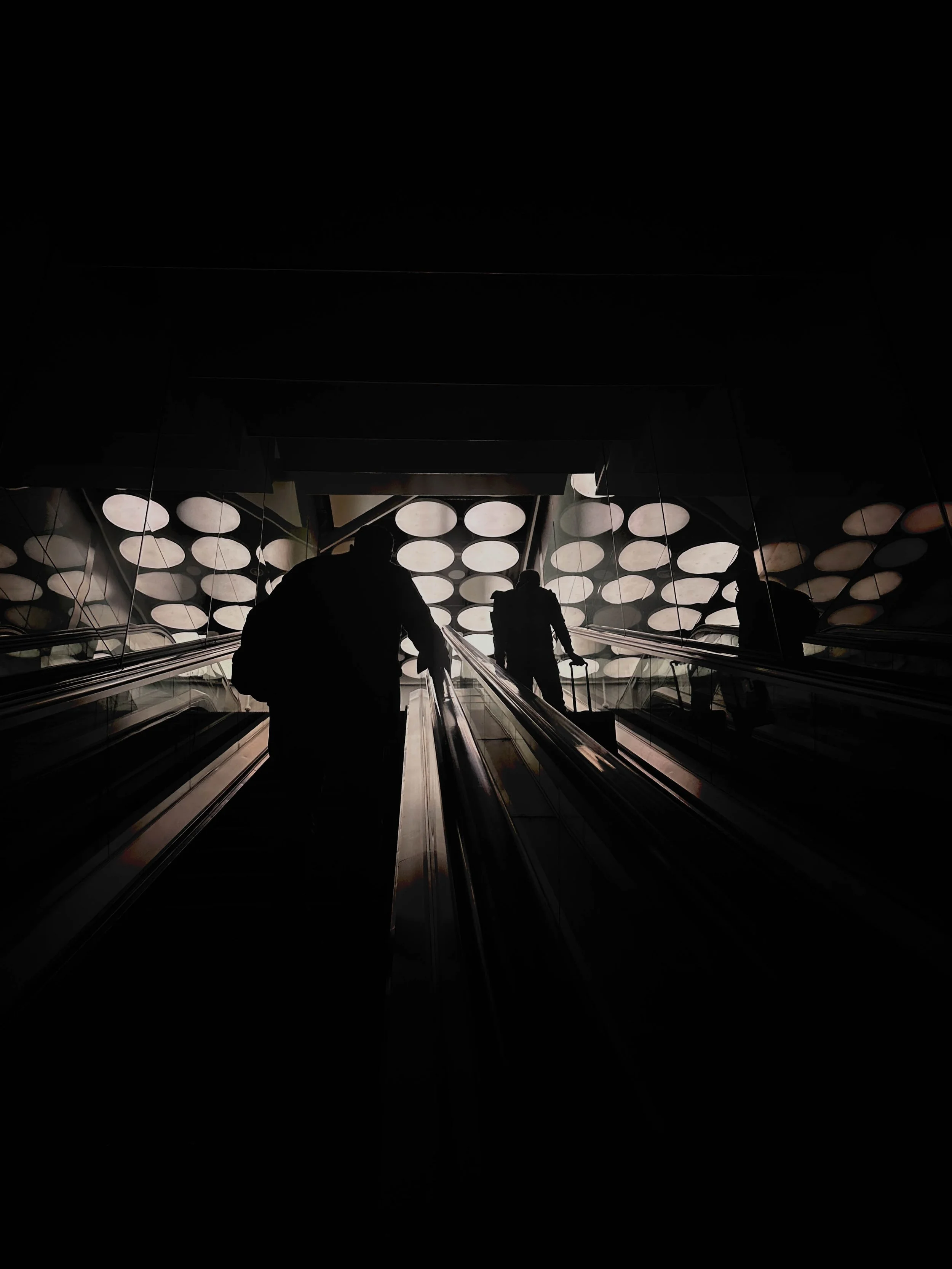 Silhouette di due persone sulla scale mobile in un edificio moderno con soffitto illuminato da cerchi di luce.