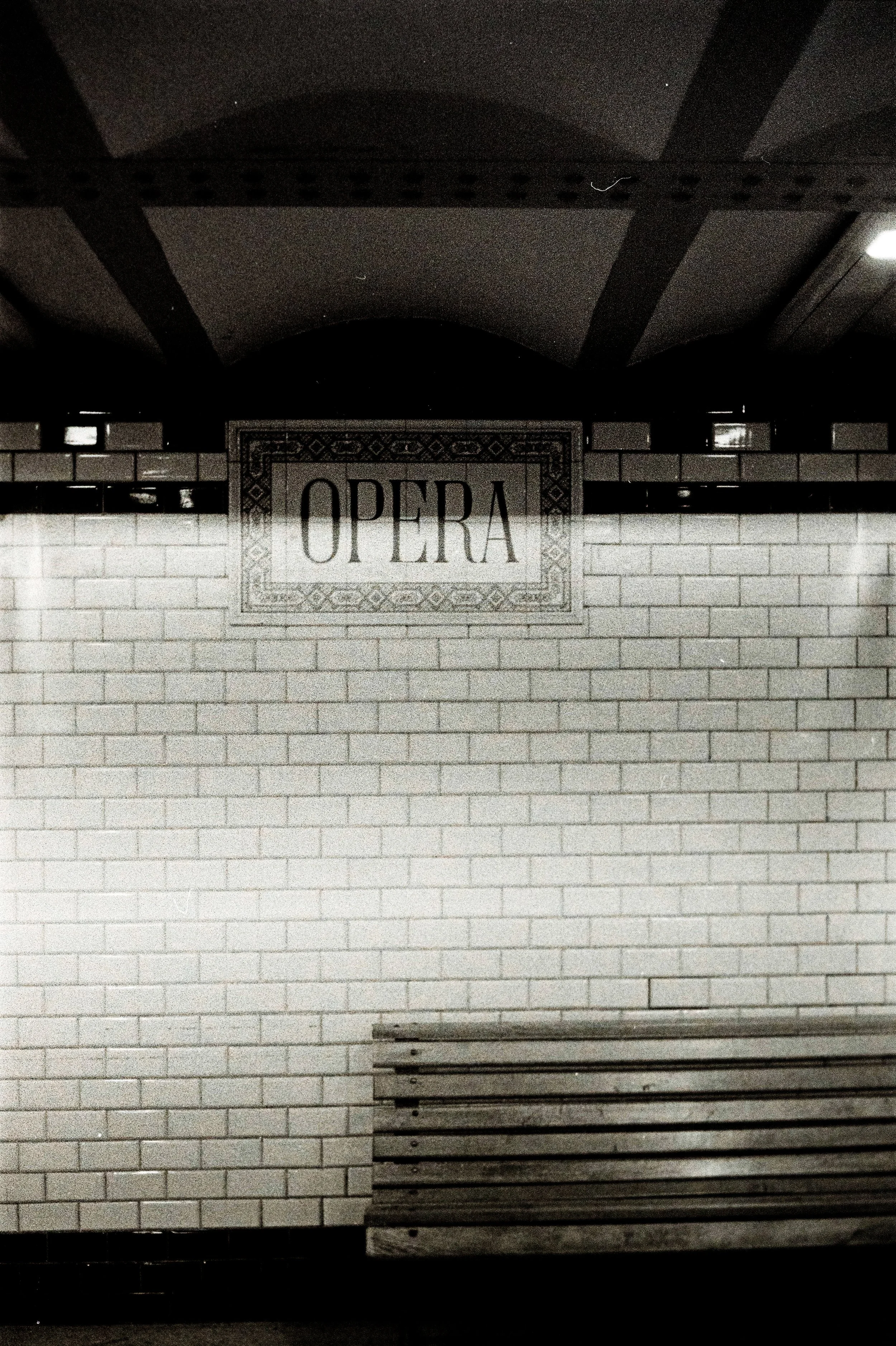 Segnale con la scritta 'OPERA' sopra un muro con mattonelle bianche e una panchina in basso.