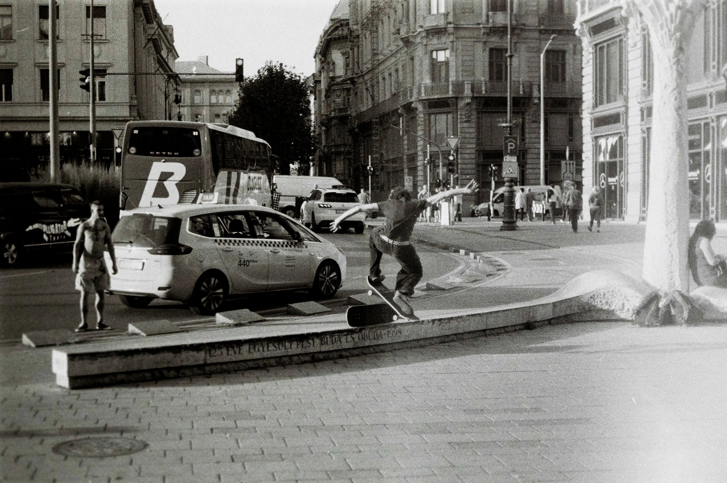 Un giovane skateboarder che esegue una manovra su una pista di skateboard in una città, con auto e autobus in background e edifici storici.