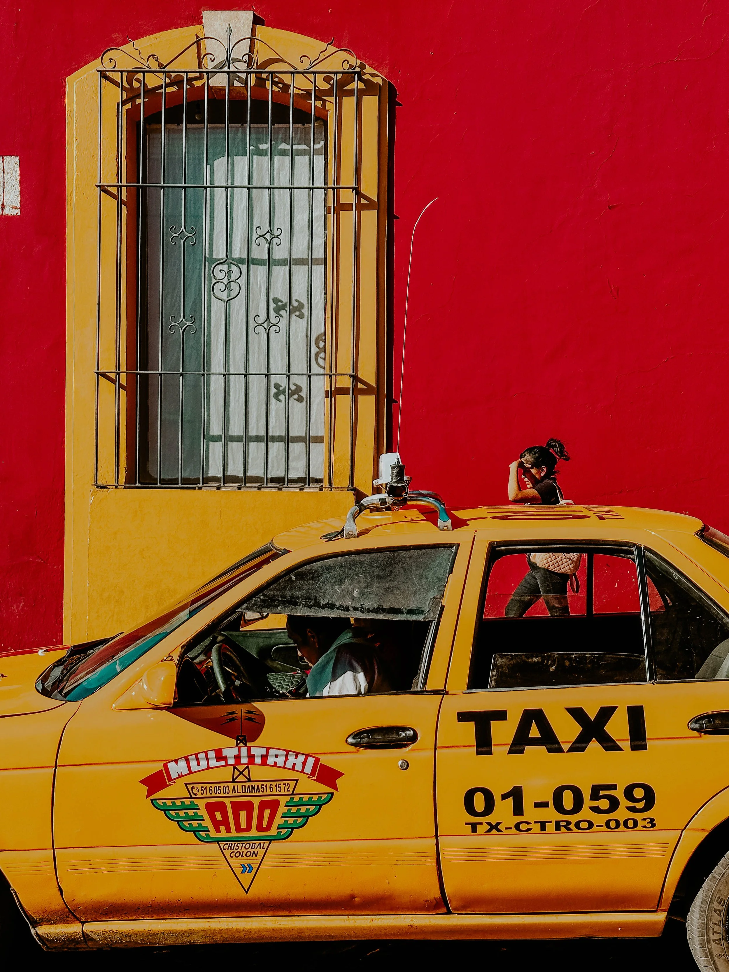 Auto taxi gialla parcheggiata davanti a un edificio con pareti rosse e gialle, una persona seduta sulla parte superiore e una donna che cammina davanti