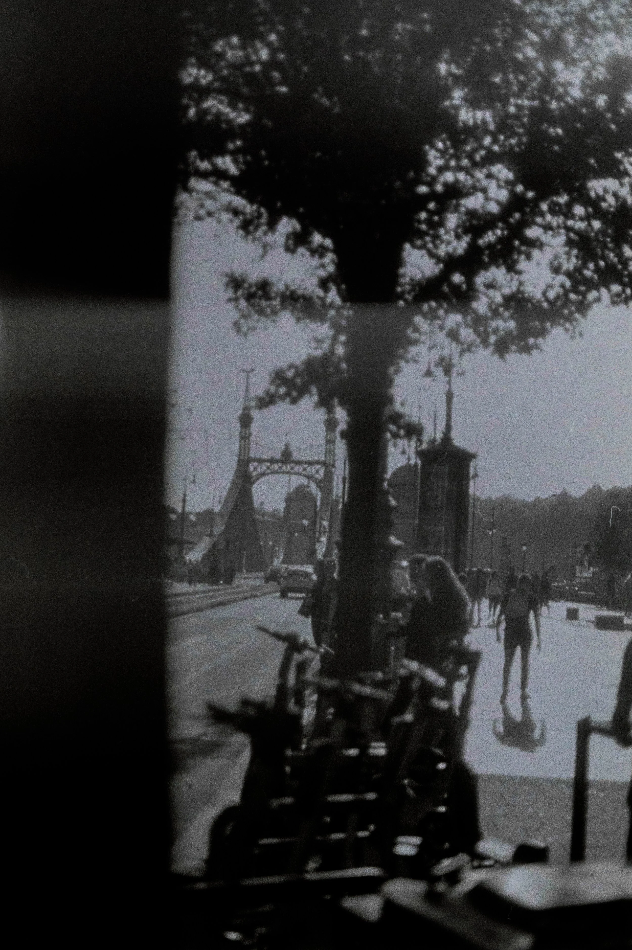 Scena cittadina in bianco e nero con un albero, biciclette in primo piano, persone che passeggiano, e un architettura con torri e ponti sullo sfondo.