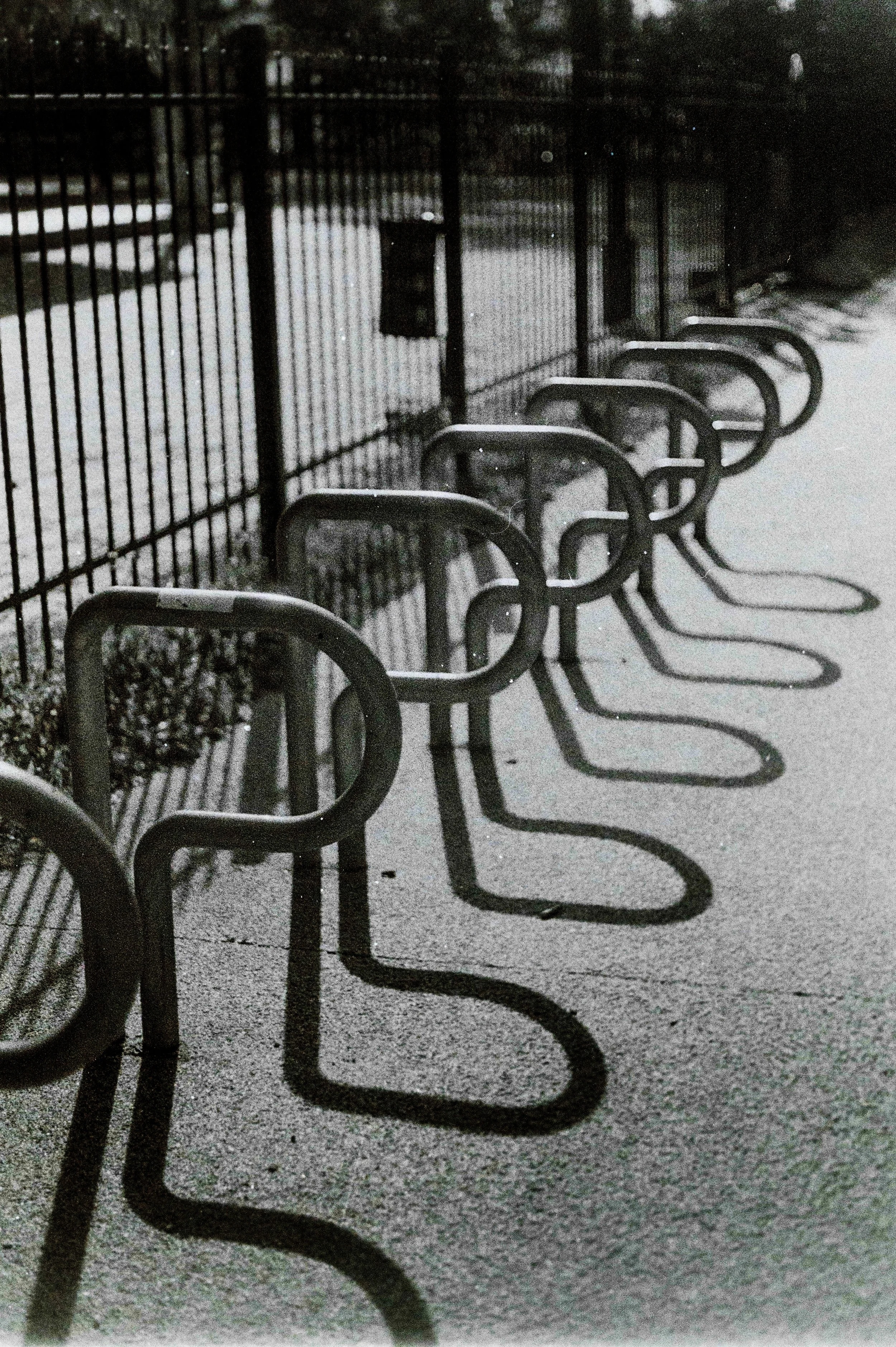 Cinque panchine di metallo senza persone, che proiettano ombre lunghe sul marciapiede, vicino a una recinzione di ferro in un'area all'aperto al crepuscolo.