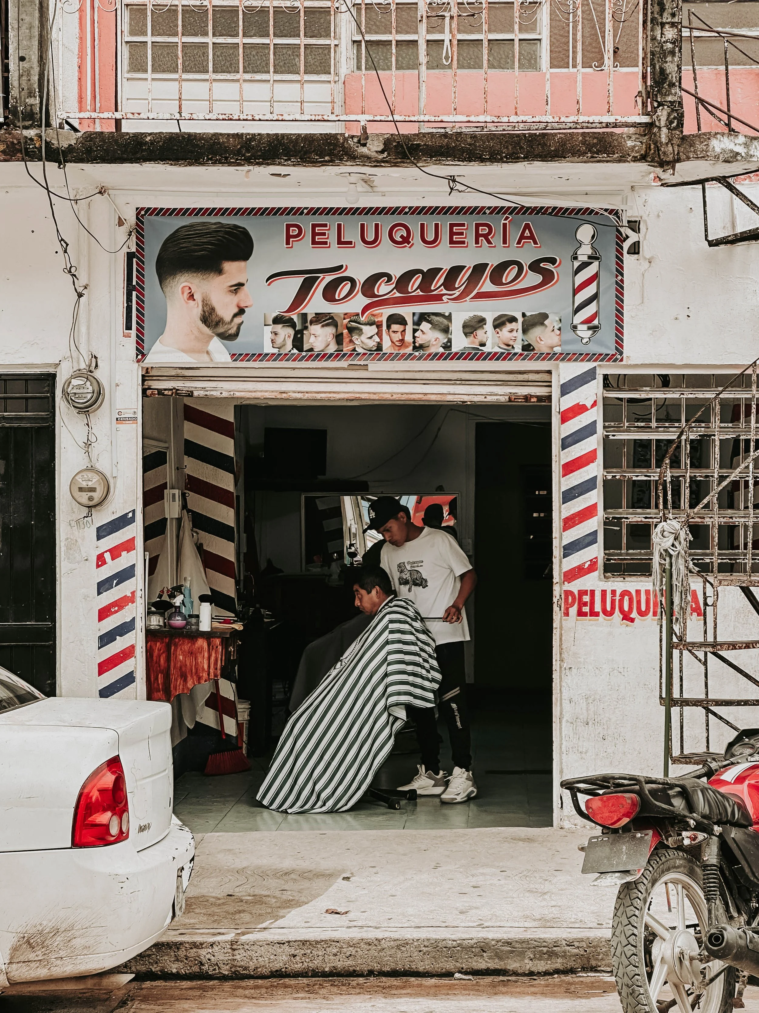Un barbiere sta tagliando i capelli a un cliente all'interno di un negozio di barbiere chiamato 'Peluquería Tocayos'. L'insegna del negozio mostra varie immagini di uomini con diversi tagli di capelli.