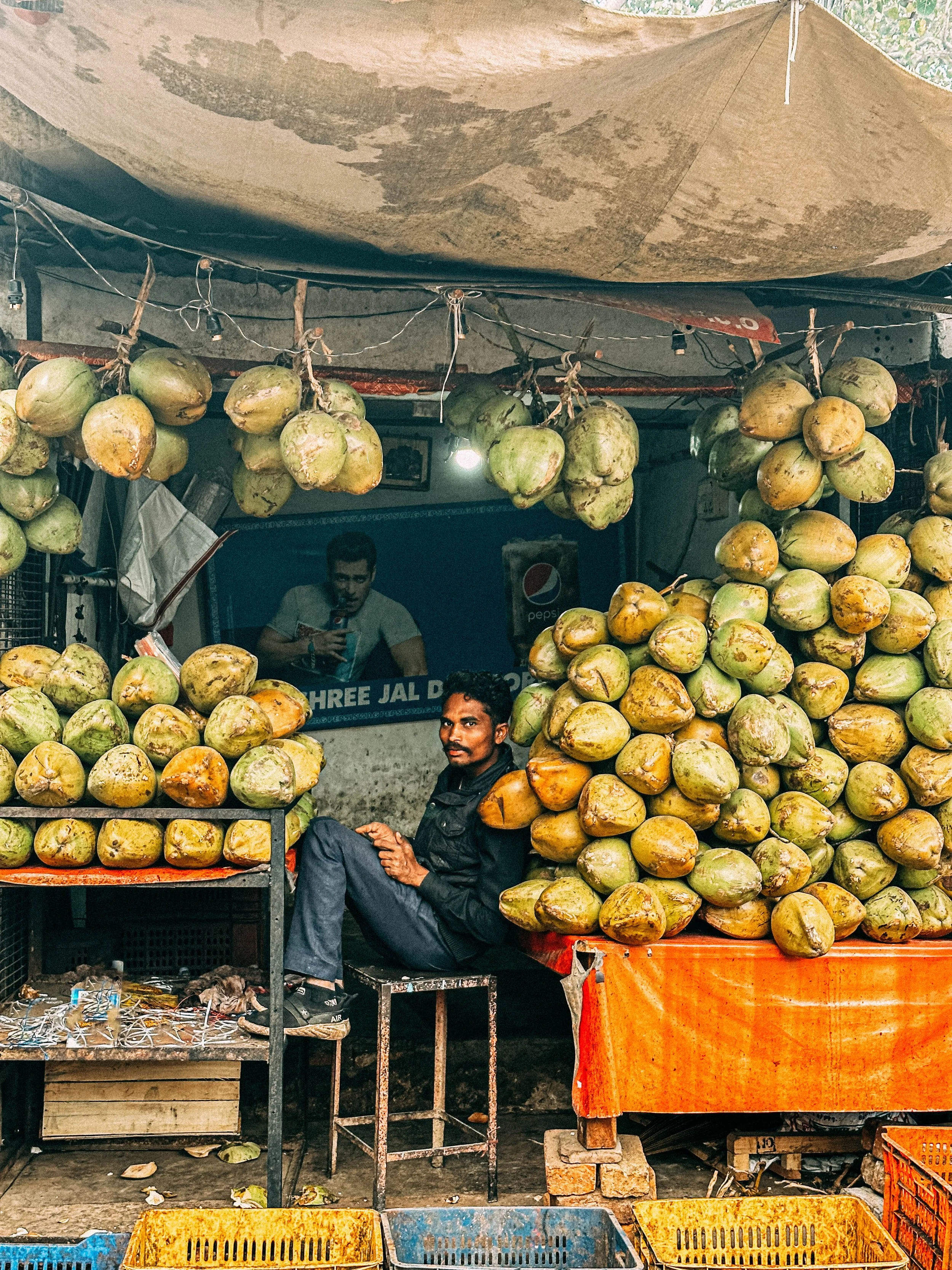 Un uomo seduto davanti a una bancarella di cocchi in un mercato, con cocchi impilati e appesi sopra. Sullo sfondo, un poster con una persona che beve una bevanda.