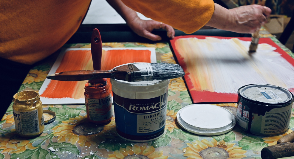 Una persona dipinge con acquerelli su carta, con vari contenitori di colori e pennelli sul tavolo decorato con fiori.