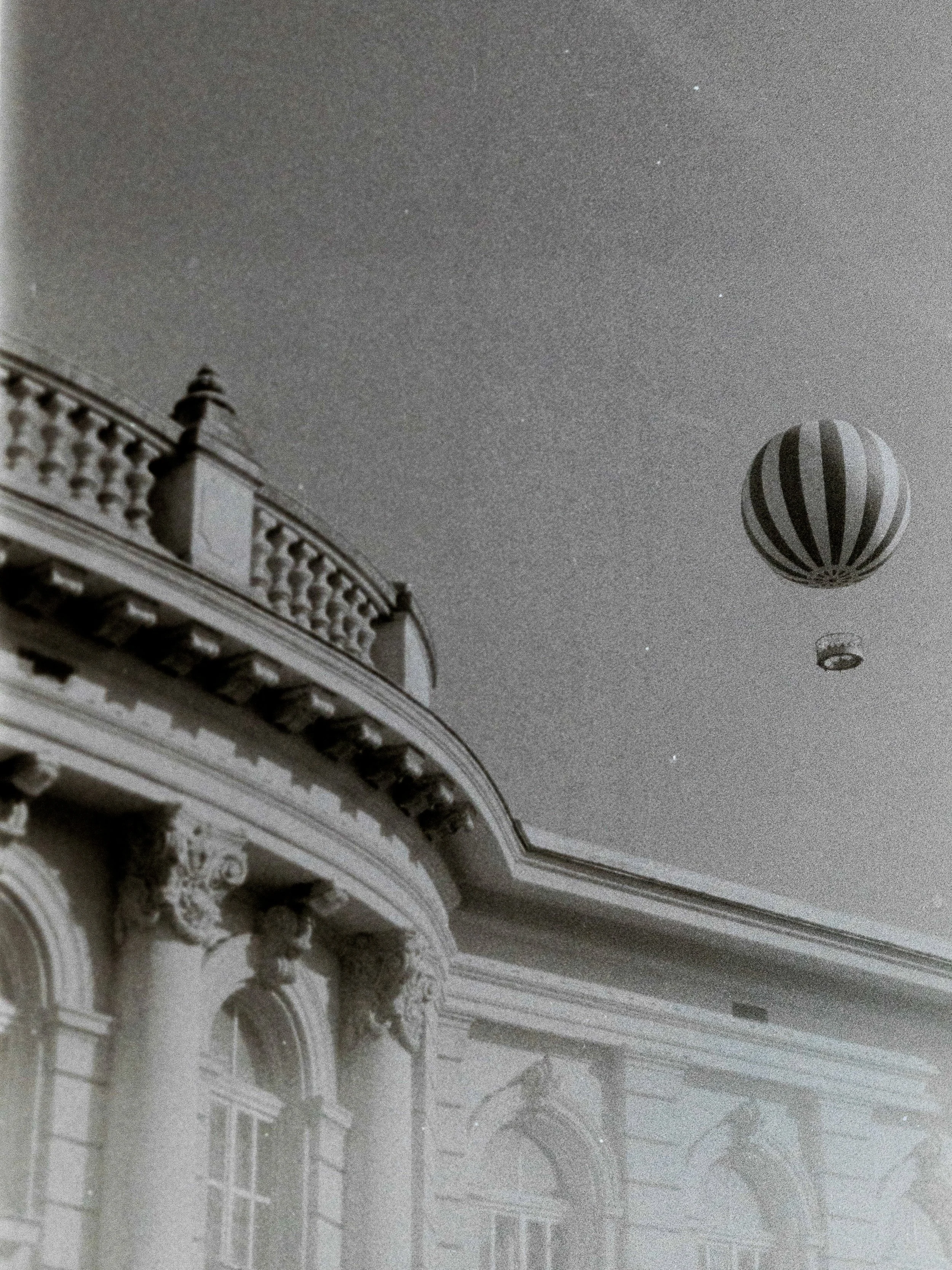 Immagine di un soffitto decorato con colonne e decorazioni classiche, con palloni aerostatici e un palloncino più piccolo che galleggiano nel cielo sopra un edificio storico.