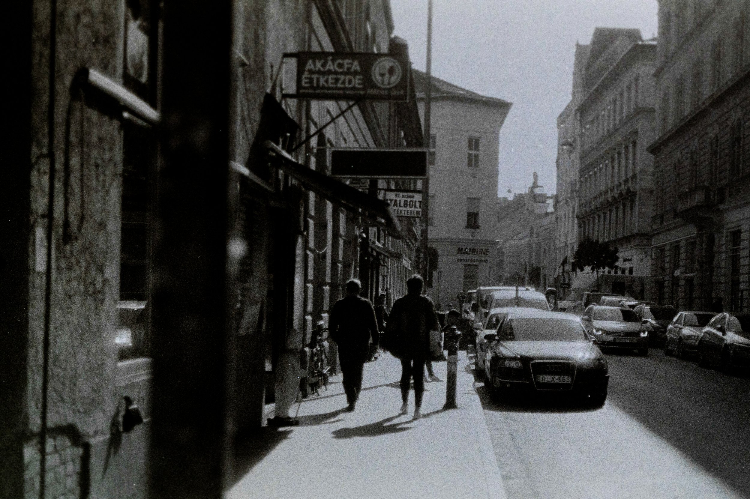 Una strada urbana con edifici alti, persone che camminano sul marciapiede e auto parcheggiate lungo la strada.