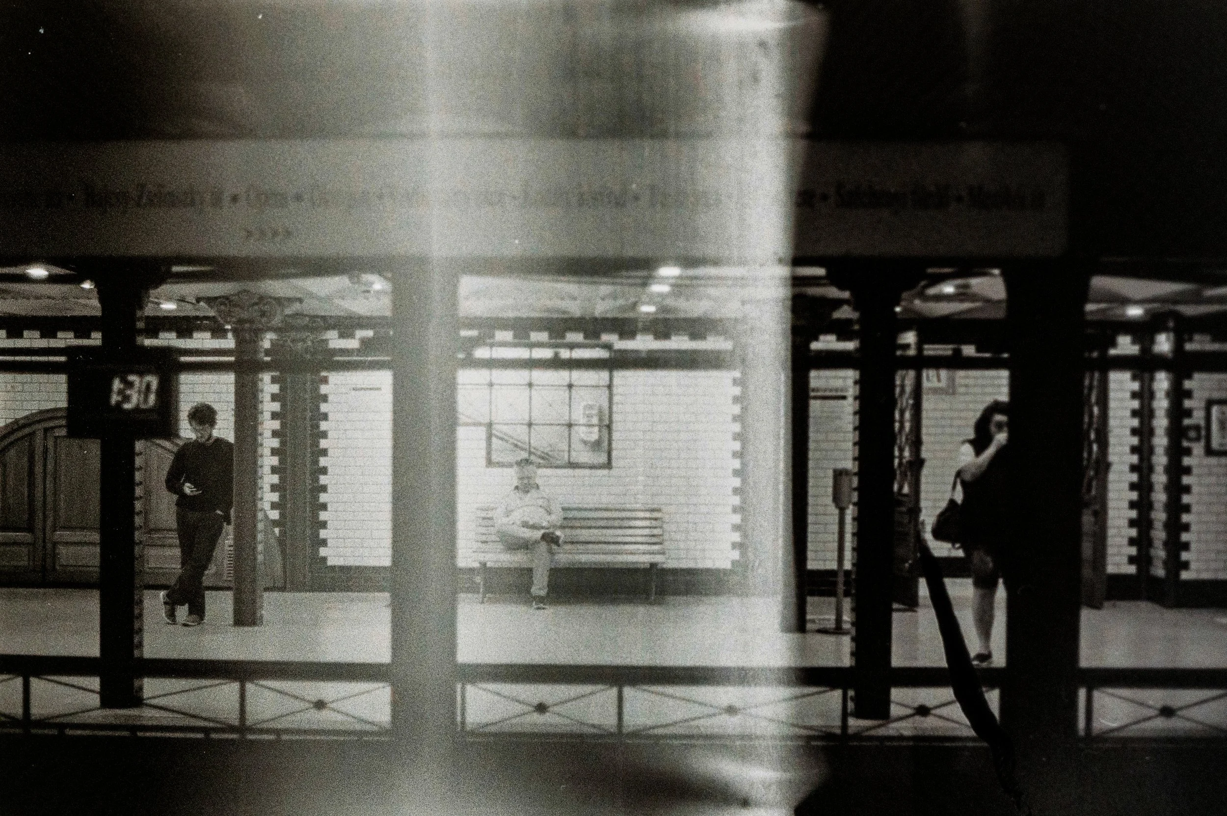 Interno di una stazione ferroviaria con persone che aspettano, pareti rivestite di piastrelle bianche, un tavolo e un canestro da basket sullo sfondo.