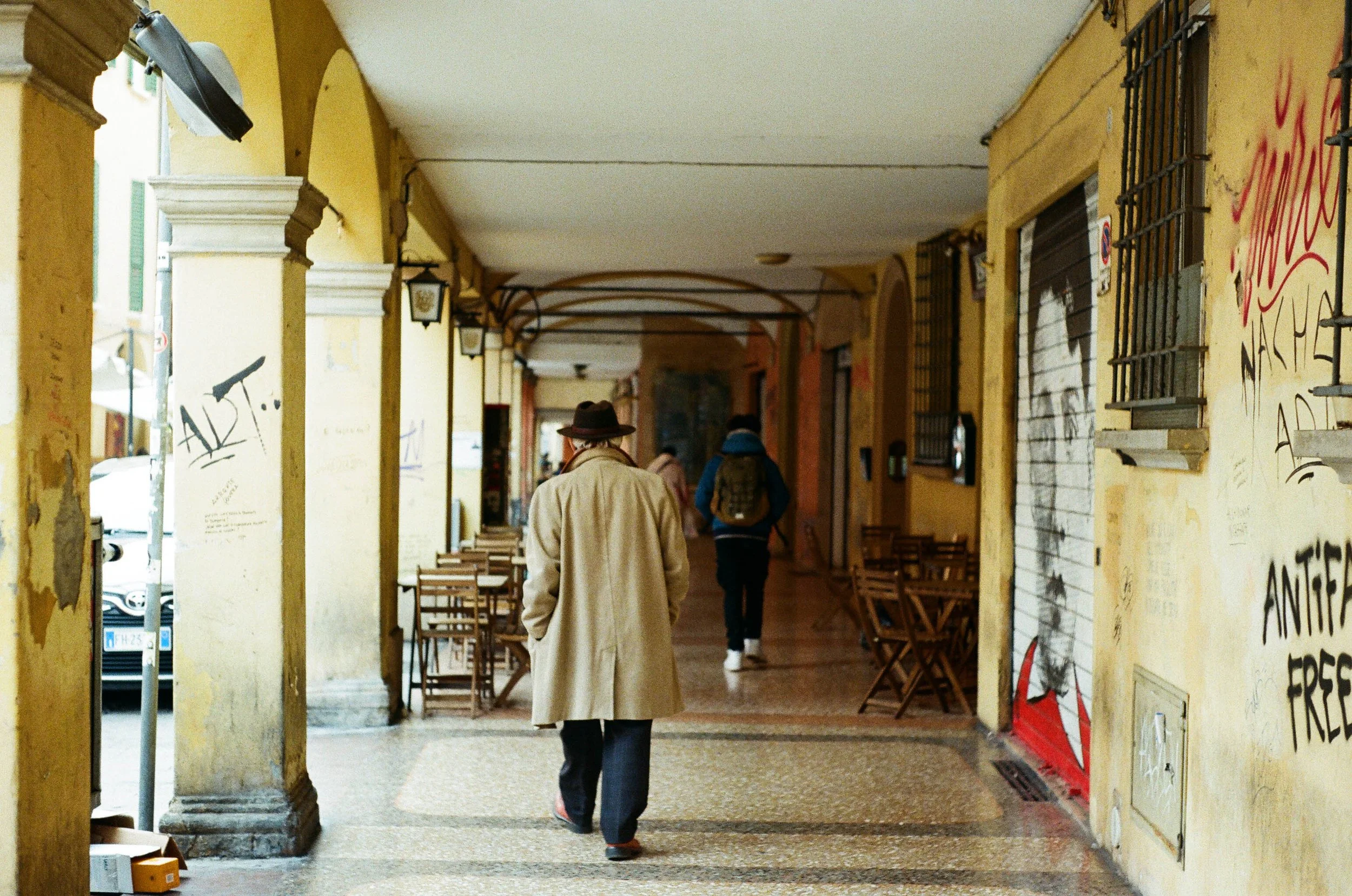 Passeggiata sotto un portico con pareti gialle e graffiti, con tavoli e sedie di legno lungo il percorso, persone che camminano, uno indossa un cappotto beige e un cappello, altri con uno zaino.
